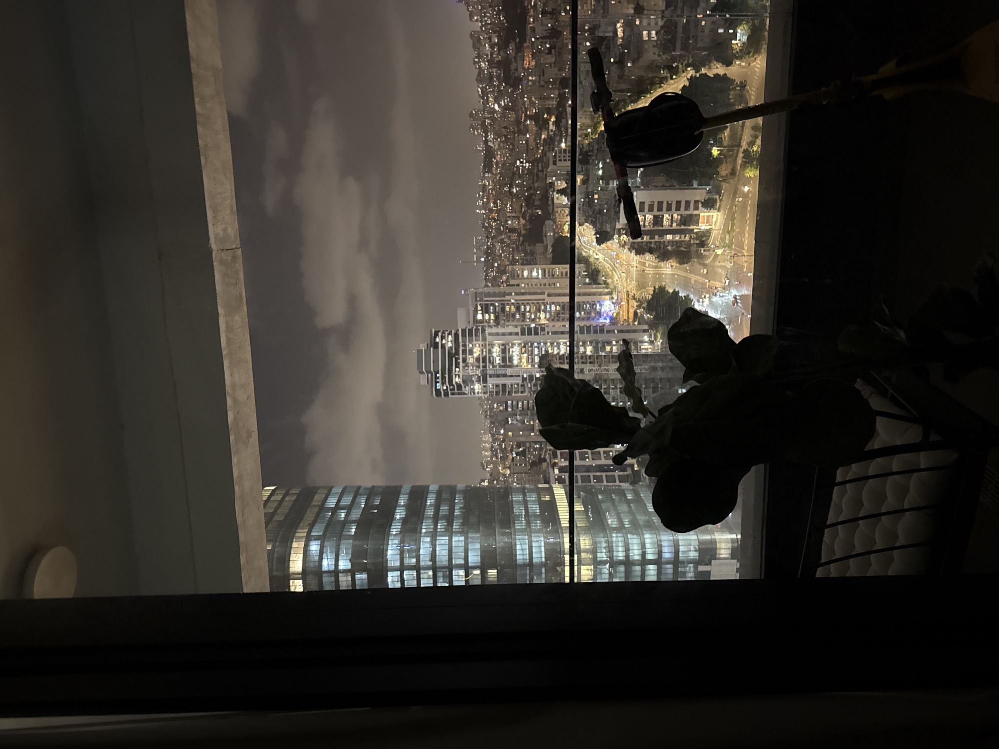 The image shows a view of Tel Aviv, Israel, at night, likely seen from a high-rise building. The cityscape is illuminated with numerous lights. Notable structures include modern high-rise buildings with lit windows. The sky above is overcast with clouds. Silhouetted objects in the foreground indicate the view is from inside a room, possibly a plant and other obscured items.