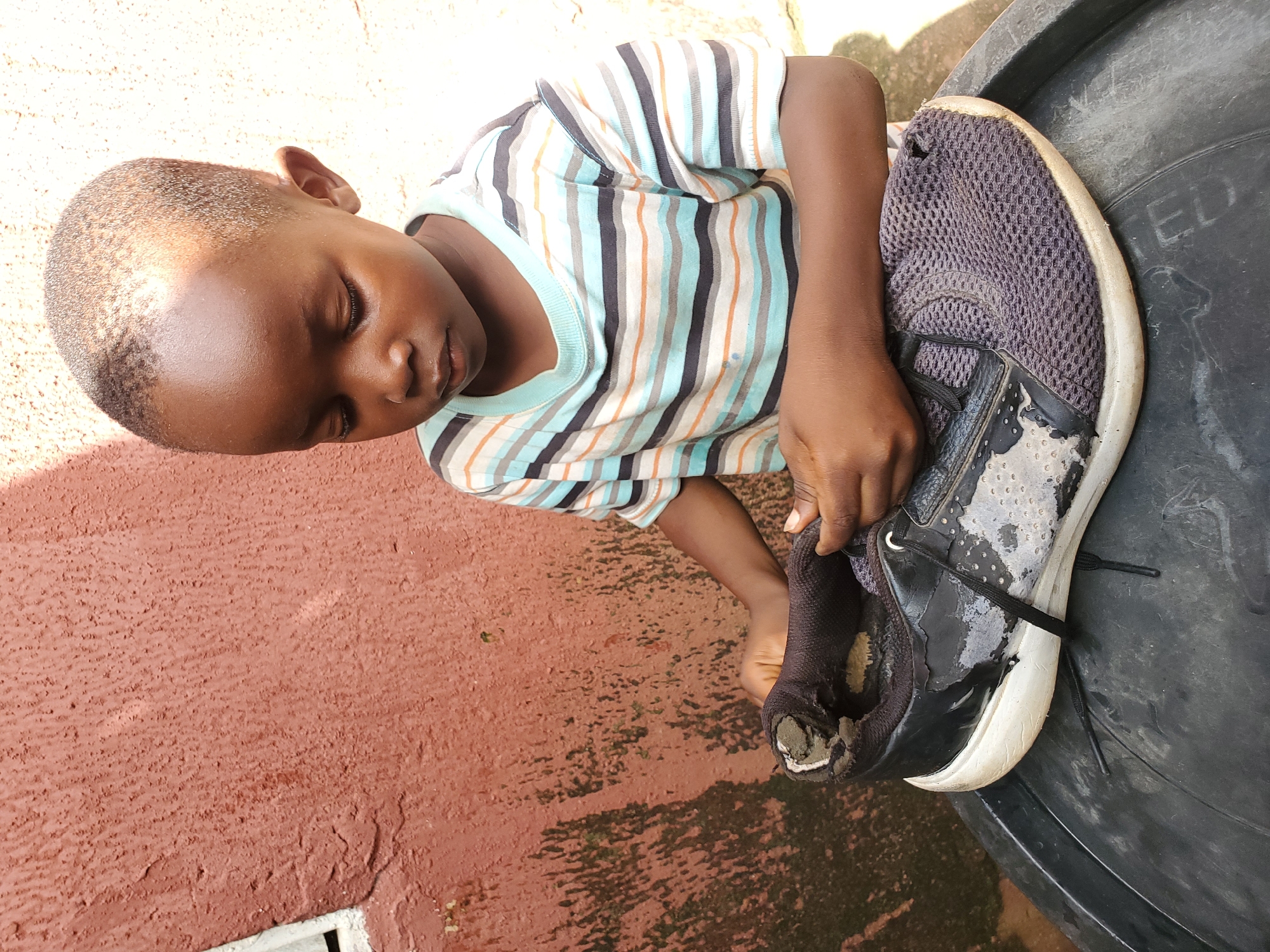 A young boy, approximately four years old, is depicted outdoors in Jos, Nigeria, with his face turned downwards and his eyes closed. He is wearing a horizontally striped t-shirt with bands of blue, white, grey, and orange. The boy is seated on a large, black, round object, possibly a plastic tub. In his hands, he is holding a worn and damaged black sneaker with a white sole. The sneaker appears to be old, with fraying fabric and peeling material on the black upper. The background consists of a textured, reddish-brown wall with patches of dark, possibly mossy, growth and a small white rectangular object, likely a window frame. Sunlight casts shadows on the wall and the boy's head.