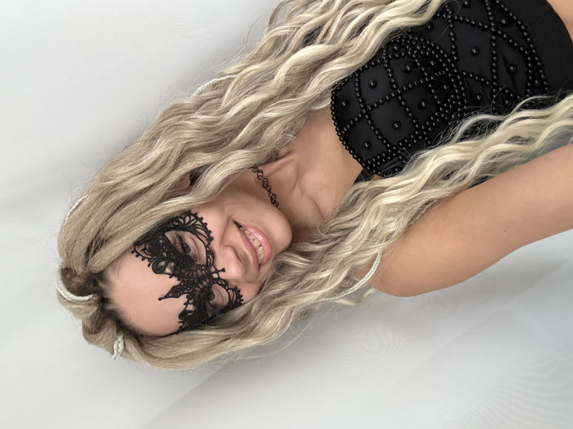 A woman with long, blonde, wavy hair is wearing a black, lacy mask covering part of her face. She is also wearing a black top with a studded pattern and a black choker. The background is a plain white surface. The image does not depict Rusanovo, Russia.