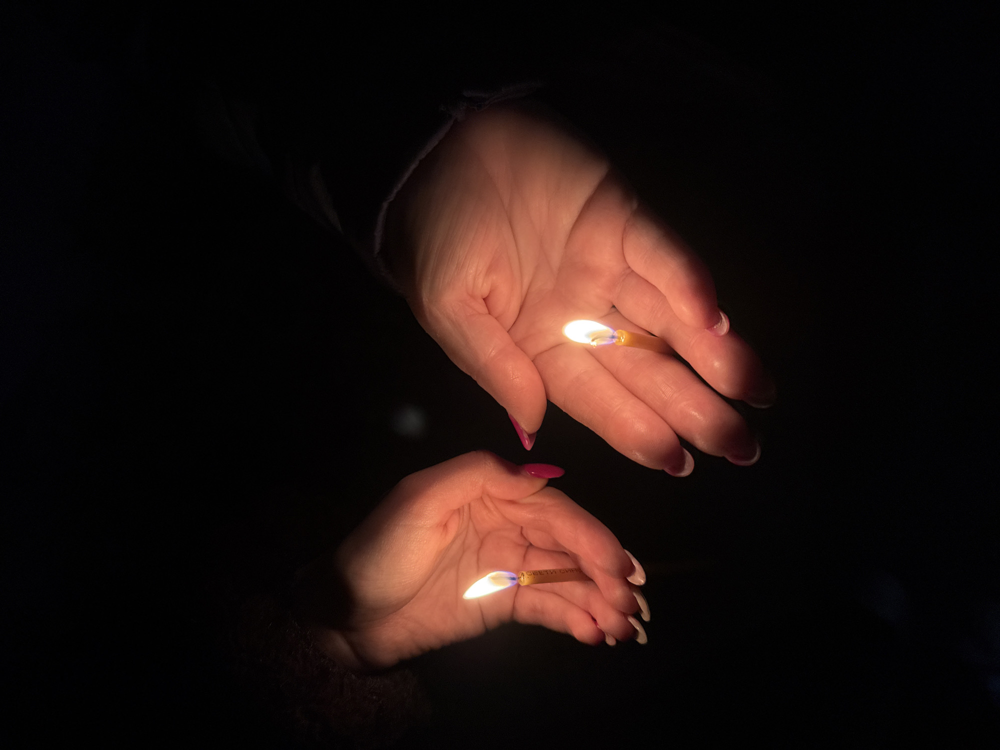 Two hands are illuminated in the dark, each holding a lit cigarette. The hands are pale and adorned with manicured nails, one set with pink polish and the other with white. The top hand is slightly angled downward towards the bottom hand, which appears to be reflecting in a dark, reflective surface. The setting is completely dark save for the light cast by the burning cigarettes, and no features indicate the scene is in Lukovit, Bulgaria.