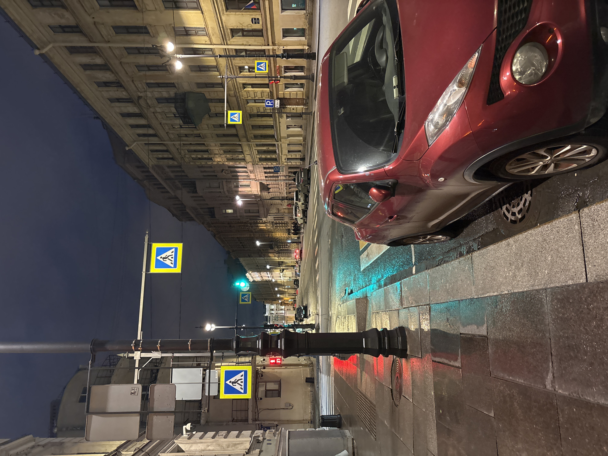 A street view in Saint Petersburg, Russia, at night. A red car is parked on the sidewalk. Crosswalk signs are visible with pedestrians depicted in blue on a yellow background. Traffic lights show red and green signals. The architecture consists of multiple-story buildings with evenly spaced windows. The street and sidewalks are wet. Dim lighting creates reflections.