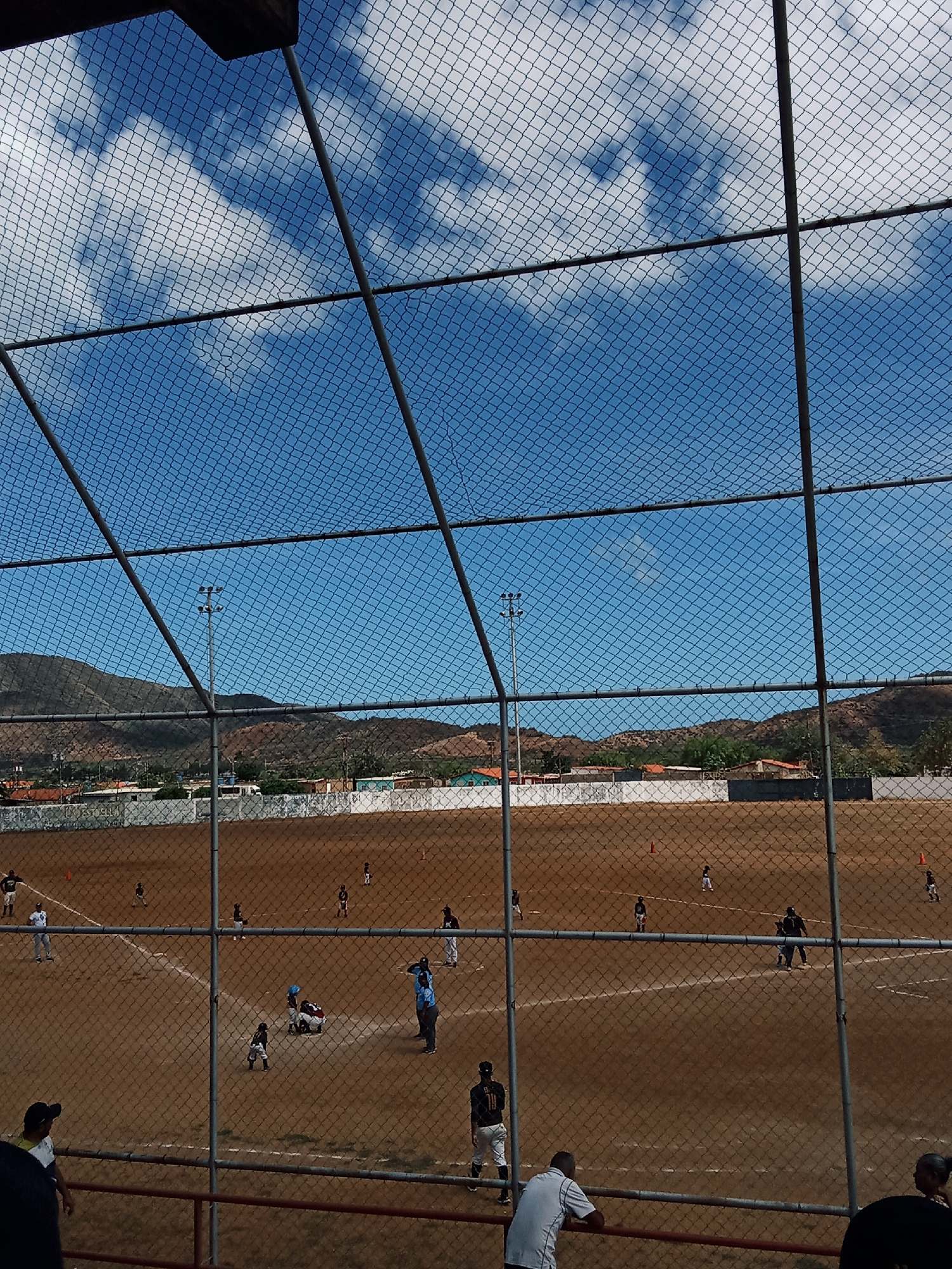 The image depicts a baseball game in progress, viewed through a chain-link fence. The field is dirt, and players are positioned in various roles. A group of players appears to be surrounding home plate. Other players are in the field. Several spectators are visible in the foreground. In the background, low mountains, buildings, and a clear blue sky are visible. Based on the presence of the mountains, the image likely portrays a location in or near the city of Juan Griego, Venezuela. There are metal light posts on the field.