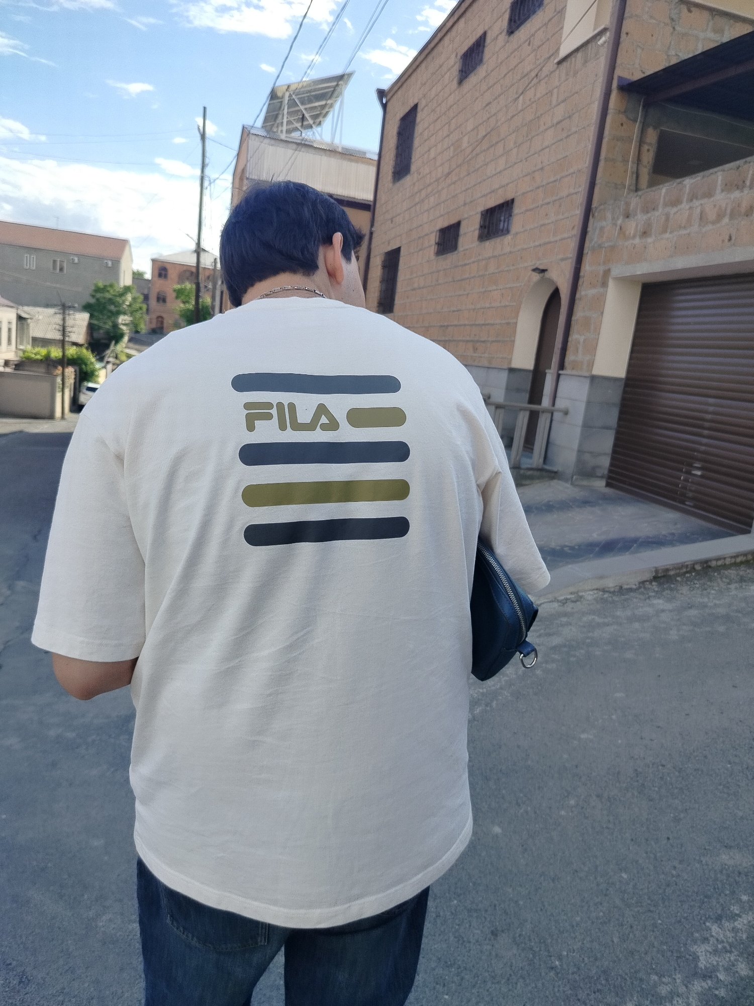 A person is standing on a street in Yerevan, Armenia, facing away from the camera. They are wearing a cream-colored T-shirt with a horizontal striped FILA logo on the back and blue jeans. A dark blue or black pouch hangs at their right side. The street is paved, and there are buildings on either side. The buildings appear to be constructed of light brown stone or brick. The sky is partly cloudy. There are utility poles visible in the distance.