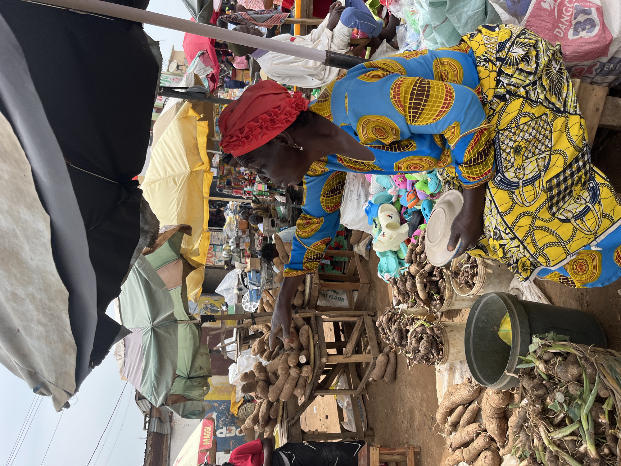 A woman in a blue and yellow dress and red headscarf sits at a market stall in Rafin Par, Nigeria, surrounded by produce. She is gesturing with her right hand, possibly towards a pile of yams. The stall is covered by a black umbrella and features wooden tables laden with yams and other root vegetables. Several buckets and bags are also visible, along with colorful sandals displayed in the foreground. Other market-goers and stalls are present in the background, indicating a bustling marketplace. The scene appears to be during daylight, with an overcast sky.
