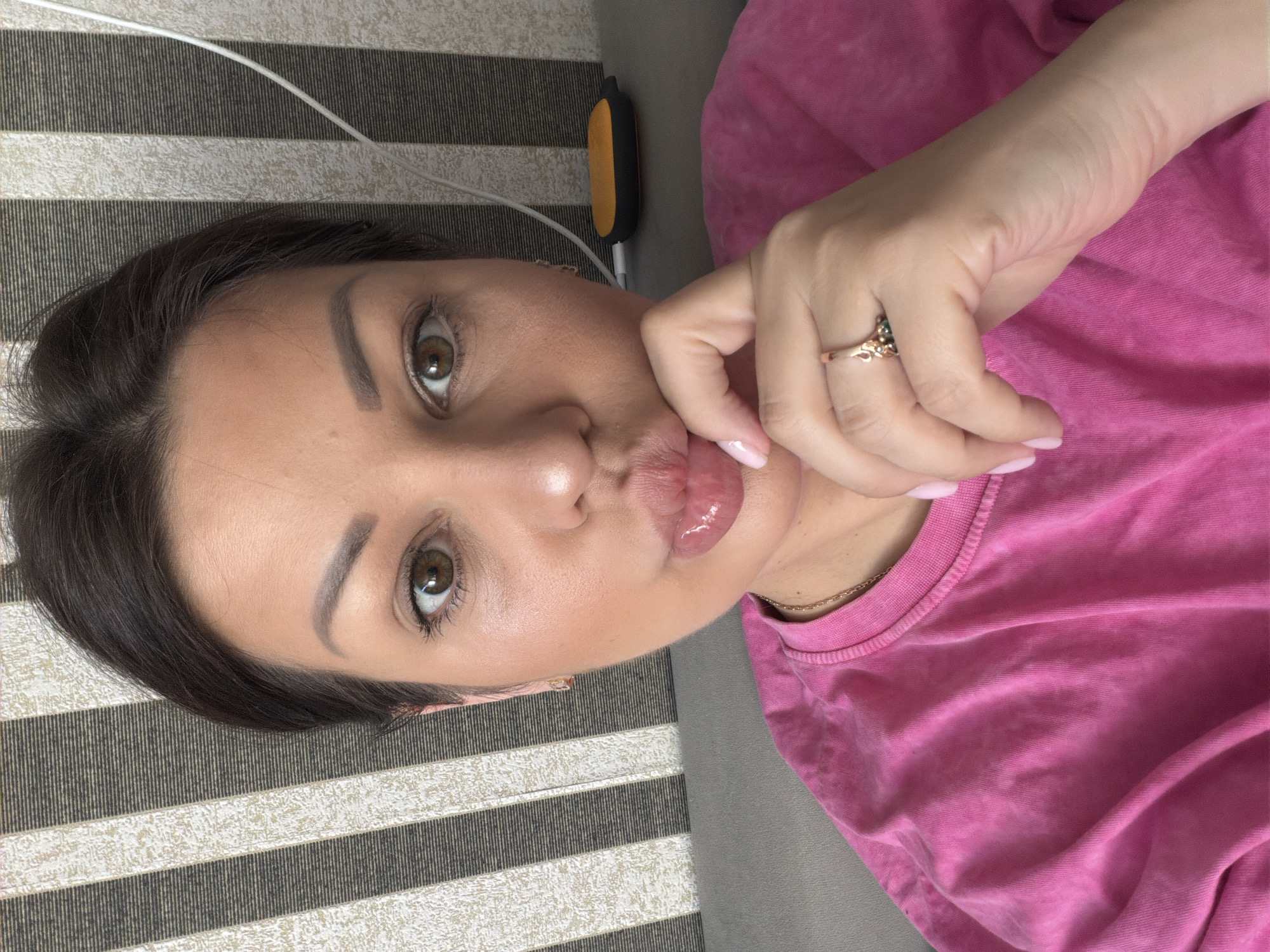 The image shows a close-up selfie of a woman with dark hair and brown eyes. She is wearing a bright pink t-shirt and has her hand touching her lips. A ring with green stones is visible on her right hand. In the background, there is a striped wall and a grey couch. A yellow and black object and a white cord are also visible in the background. There is no indication of the city of Sochi, Russia in the image.