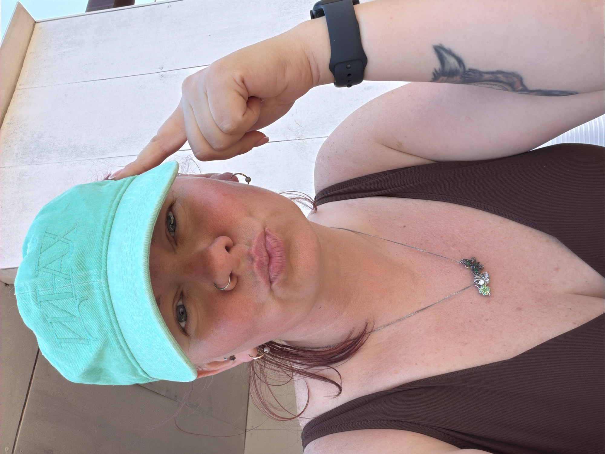 A woman is taking a selfie outdoors. She is wearing a mint green baseball cap, a brown top, and a black smartwatch. She has a tattoo on her arm. Her finger is pointed towards the brim of her cap. She has multiple piercings in her ear and a nose ring. She is also wearing a necklace. The background appears to be a light-colored wall with horizontal lines. There is no information available to identify the location in Chaikovsky, Russia.