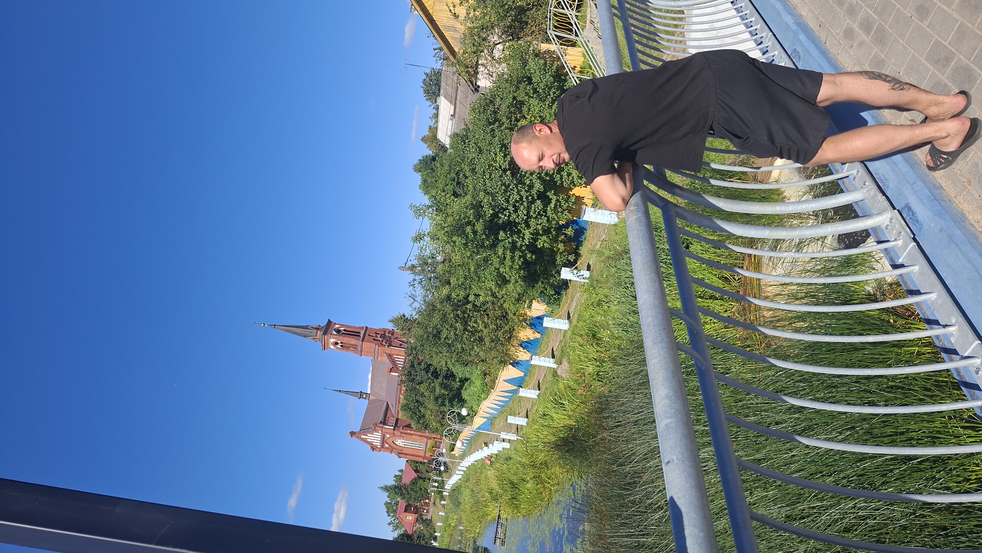 A man stands on a bridge, leaning on the railing and looking towards a church with a tall steeple. The church is made of red brick and has a Gothic architectural style. It is situated on a hill surrounded by trees. A path with lampposts leads up to the church from a grassy area. The bridge has a metal railing and a blue base. The man is wearing a black t-shirt and black shorts. The sky is clear and blue. This scene is located in the city of Pastavy, Belarus, and depicts the Church of the Transfiguration of Jesus.