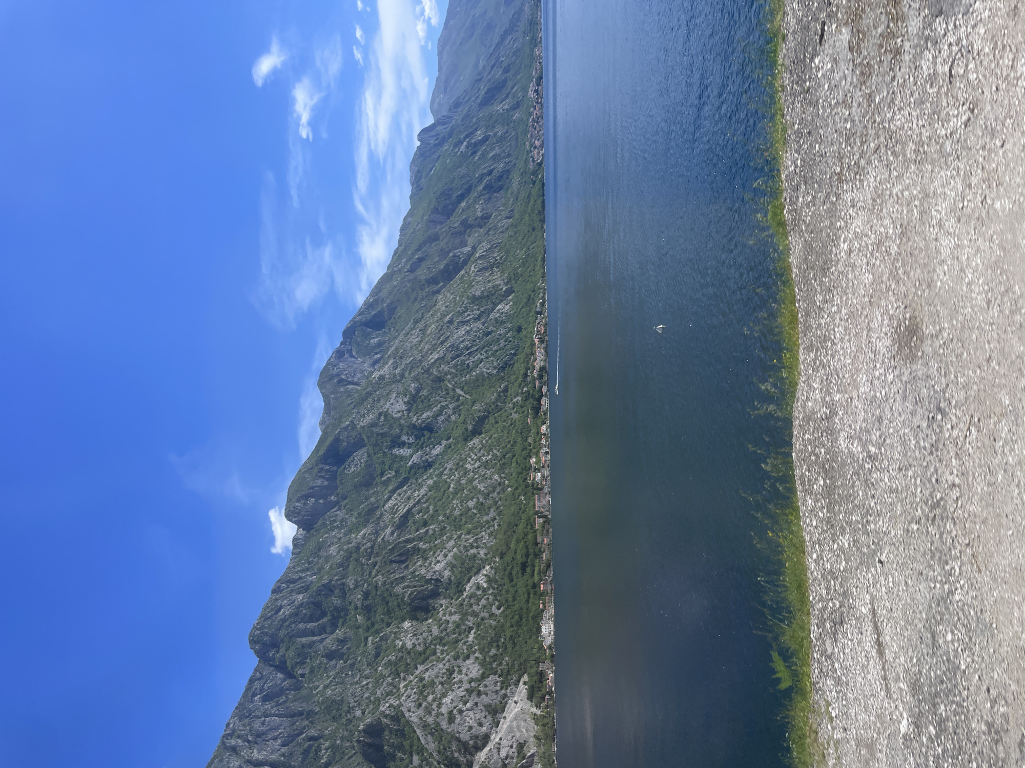 The image depicts a scene in Donji Orahovac, Montenegro. A steep, rocky mountain covered with sparse vegetation rises on the left side of the frame. A settlement with buildings is nestled along the base of the mountain where it meets the water. The body of water, likely the Bay of Kotor, occupies the central portion of the image. It's calm and dark, reflecting the light from the sky. On the right, a gray, textured surface, potentially a concrete pier or a rocky shore, runs along the water's edge. The sky above the mountain is blue with scattered white clouds. A small boat appears to be sailing on the water.