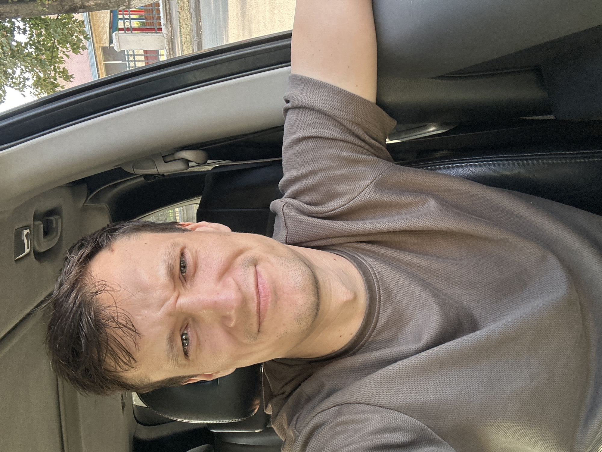 A man is captured in a selfie taken from inside a car, with his face and upper torso filling the frame. He is wearing a brown, short-sleeved shirt. His right arm is resting on the car's armrest, visible on the right side of the image. The interior of the car, including a black leather seat and a gray pillar, is evident. Outside the car, a street scene unfolds, featuring a sidewalk, trees, and a building with a red and blue facade, suggestive of the city of Kamianets-Podilskyi, Ukraine, given the distinctive architecture visible in the background.