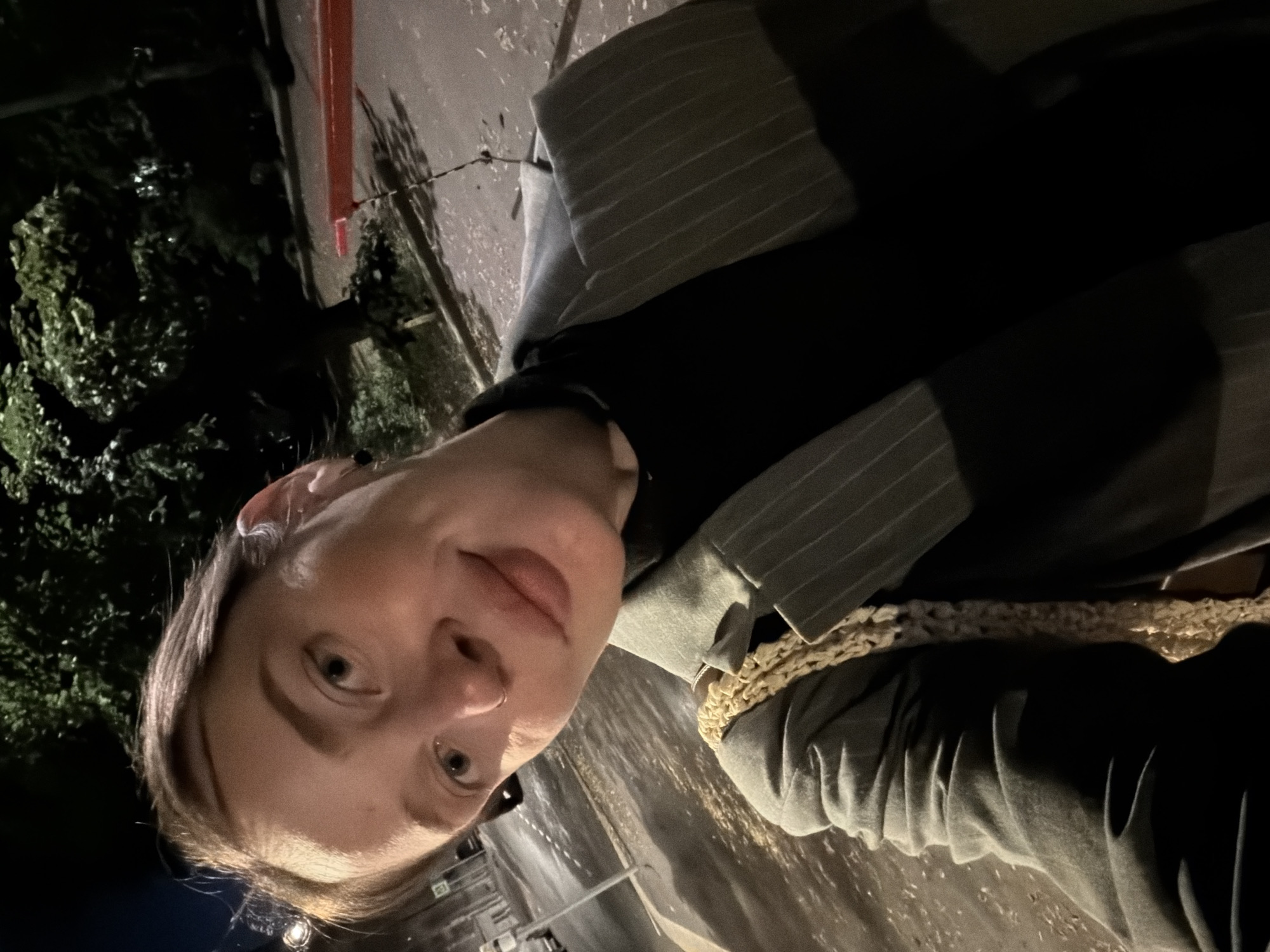 The image is a selfie taken outdoors at night, possibly in an urban environment. The subject is a young woman with light skin and light brown hair pulled back. She is wearing a grey pinstriped blazer over a black turtleneck and a cream-colored woven bag is visible on her shoulder. She has a nose ring and an ear piercing. In the background, there are trees, streetlights, and a street that appears to be wet, suggesting recent rain or it is nighttime. A red barrier is visible in the middle right of the frame. The scene appears to be in Moscow, Russia, based on the presence of Cyrillic signage on a pole in the background, which is consistent with Russian street signs.