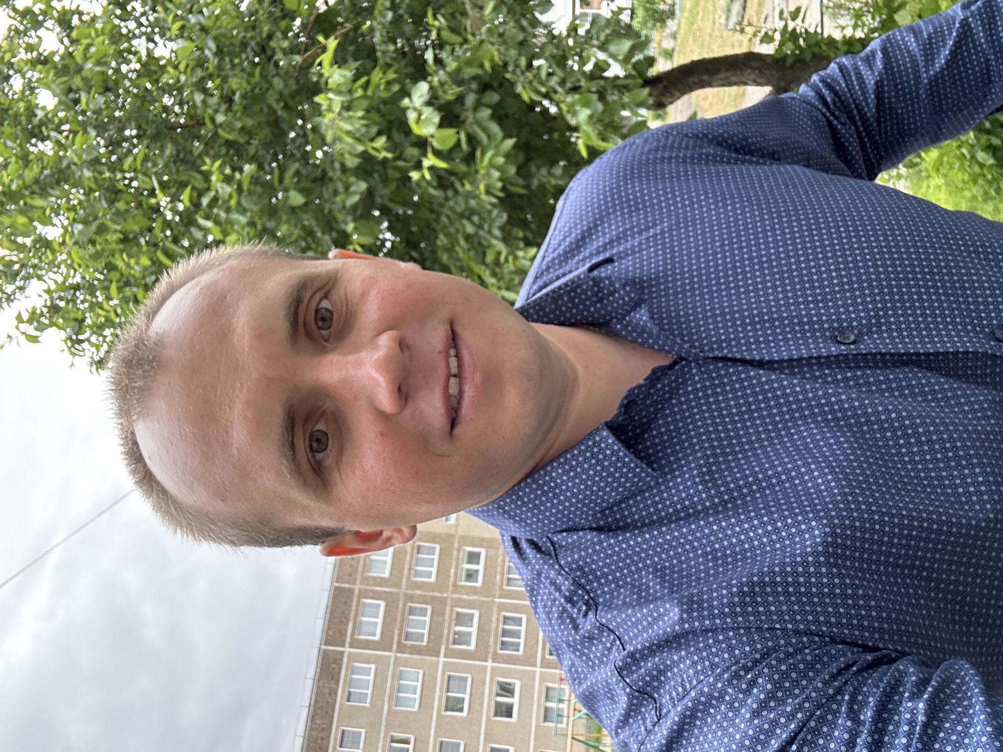 A man is taking a selfie in front of an apartment building in Chaikovsky, Russia. The man is wearing a blue, long-sleeved, patterned shirt. The building behind him is multi-storied and gray, with visible windows. A tree with green leaves is visible above and behind the man. The sky is cloudy.