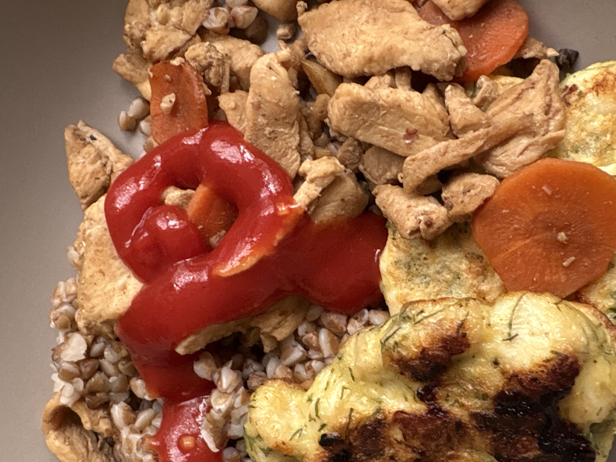 A close-up, high-angle shot captures a rustic meal served in what appears to be a light grey or beige bowl or plate. The dish features a mix of different food items, suggesting a hearty, home-cooked plate.
The primary components visible are numerous pieces of cooked, light brown meat, likely chicken, cut into irregular strips and chunks. These pieces have a somewhat textured, possibly pan-fried or baked appearance. Interspersed among the chicken are small, light brown, granular grains, consistent with cooked buckwheat groats.
Bright orange, round slices of cooked carrots are visible, adding a pop of color to the otherwise earthy tones of the chicken and buckwheat. A generous swirl of glossy, vibrant red sauce, resembling ketchup, is squeezed across the center of the dish, creating a prominent visual element.
To the bottom right, several yellowish-golden fritters or patties are present. These appear to be pan-fried, exhibiting some darker, crispy, and browned patches. Green specks, consistent with chopped dill or other herbs, are visible within the fritters, suggesting a savory flavor profile.
The food is presented compactly, with ingredients piled together. There is no discernible activity taking place beyond the presentation of the meal. The image is tightly cropped, offering no background details to indicate the time of day, weather, or specific location, although the cuisine components, particularly buckwheat and the style of fritters, could align with culinary traditions found in Saint Petersburg, Russia, as per the provided context. There is no visible text in the image.