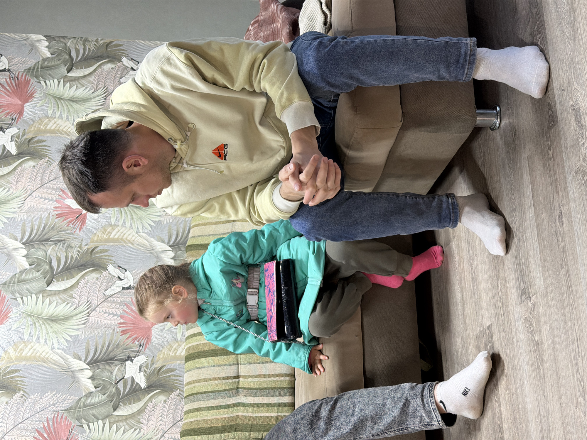 A photograph taken in Chaikovsky, Russia, depicts an indoor scene. A man wearing a light yellow hoodie and blue jeans sits on a brown couch, holding the hand of a young girl who is lying on a striped cushion. The girl is wearing a teal jacket, green pants, and bright pink socks. She is carrying a black purse with a chain strap. The background includes a wall with a leaf and bird-patterned wallpaper. The floor is light-colored wood. Another person's legs are visible in the foreground, wearing gray jeans and white socks with a Nike logo.