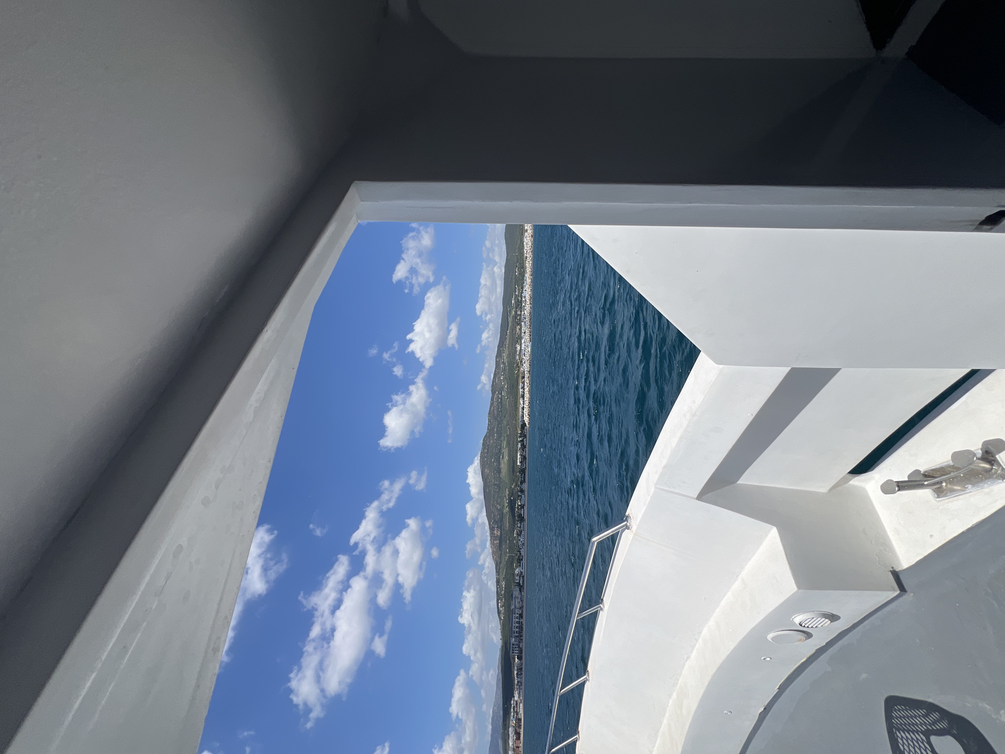 The image is taken from inside a boat, looking out through an opening. The outside scene consists of a blue sky with puffy white clouds, a body of water (likely the Mediterranean Sea), and a coastline with a city.
The city along the coastline is M'Diq ⵎⴹⵢⴰⵇ المضيق, Morocco, evidenced by the geography and architectural style.
The boat's interior elements, such as the white walls and part of the deck, are visible. The boat is in motion on the water.