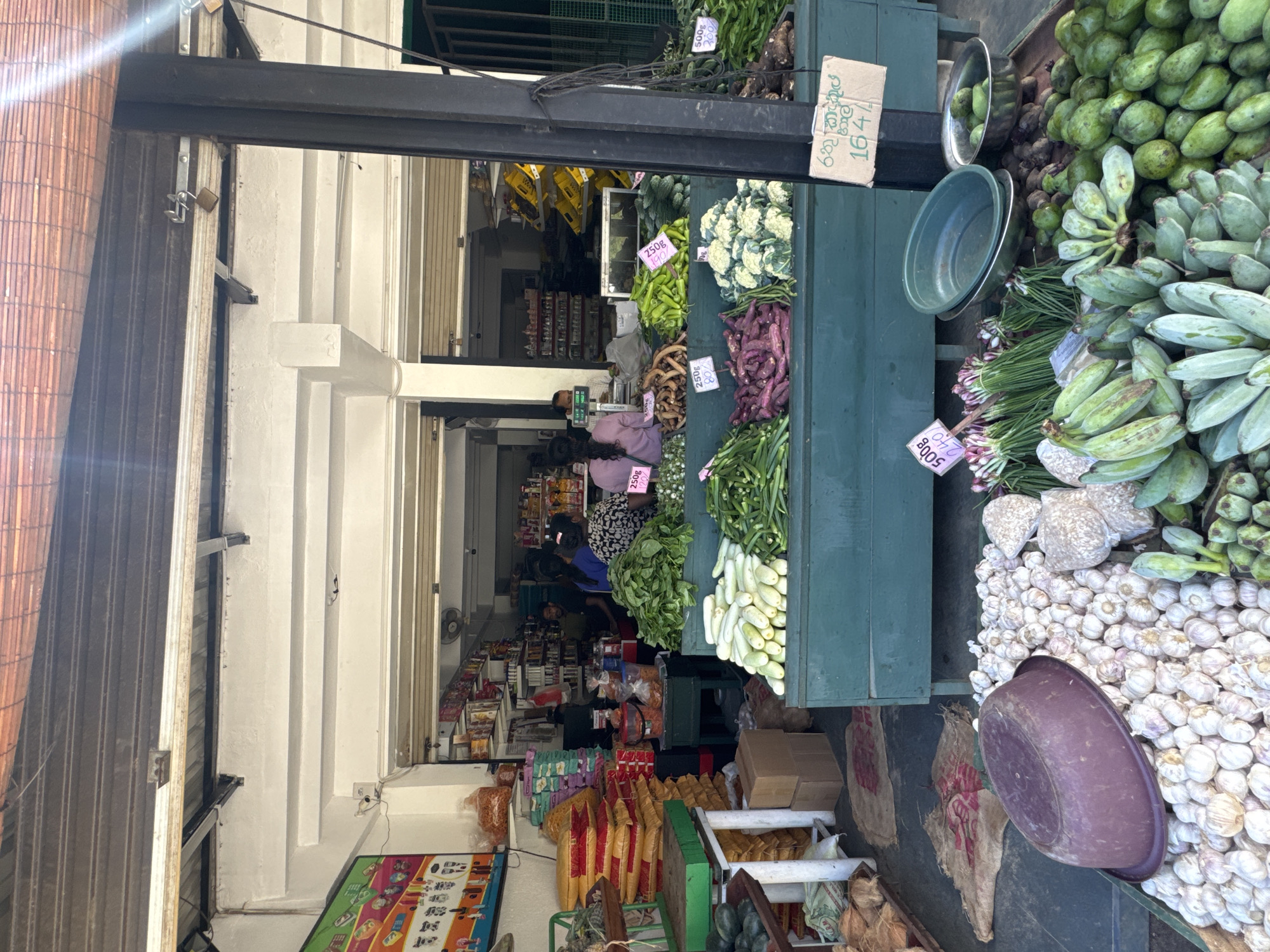 An open-front market stall in Matara, Sri Lanka, teems with fresh produce on a bright day. The scene is captured looking into the stall from under a covered area, with a metal roll-up door visible to the left.
On the right side of the image, large quantities of green bananas or plantains are piled high on a green wooden display, alongside smaller, round green fruits, possibly raw mangoes. Below these, bundles of green onions or scallions are arranged next to a substantial heap of white garlic bulbs, upon which a dark purple plastic bowl rests.
In the center, various fresh vegetables are displayed on another green wooden stall. These include green leafy vegetables, white cauliflower, long purple eggplants (brinjals), green beans, bright green chilies or peppers, and white cucumbers or gourds. Further inside the shop, a person with dark hair, wearing a purple top, is seen near a weighing scale, with another individual visible deeper within the store. The interior reveals shelves stocked with various packaged goods, suggesting a combined grocery and produce establishment.
The activity centers around the sale of fresh fruits and vegetables. The market stall appears active, with products neatly arranged for customers.
Several price tags are visible, providing notable details:
* "500g 100/-" on green leafy vegetables.
* "250g 90/-" on green chilies/peppers.
* "250g 80/-" on purple eggplants.
* "500g 240/-" on green onions/scallions.
* A cardboard sign displays "ඉඟුරු කිලෝ 164/-", which translates to "Ginger kilo 164/-".
* Another tag shows "250g 90/-" on the white cucumbers/gourds.
* A partially visible tag on green beans indicates "250g 120/-".
The prices likely refer to Sri Lankan Rupees (LKR). The overall setting conveys a bustling local market atmosphere.