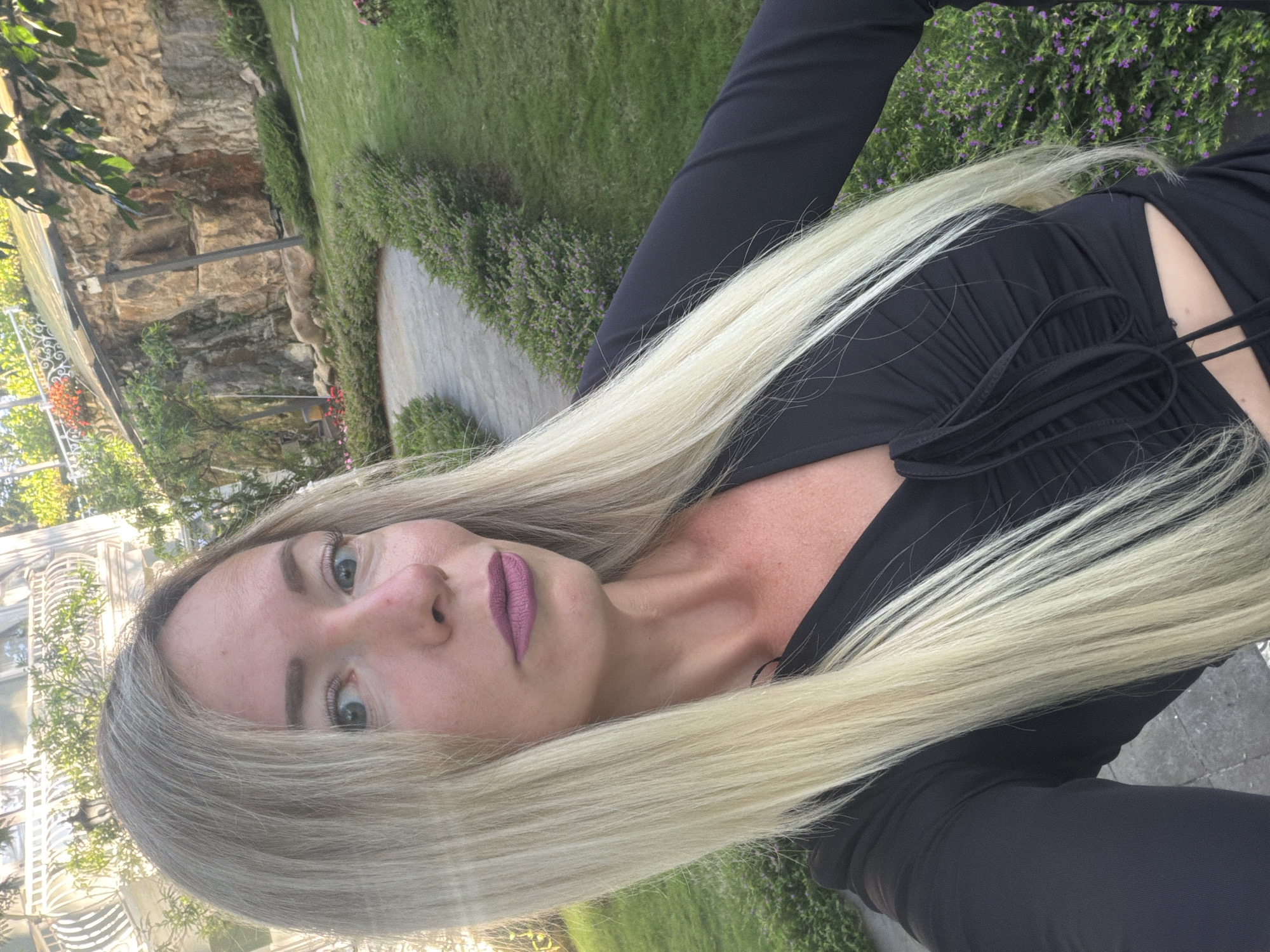 A blonde woman wearing a black dress with a cutout is taking a selfie outdoors. She has on purple lipstick and her hair is long and straight. The background appears to be a garden area with a stone wall and lush green grass. There is also a glimpse of a white building with ornate balconies. The location is identified as Xuân Trường - Đà Lạt, Vietnam.