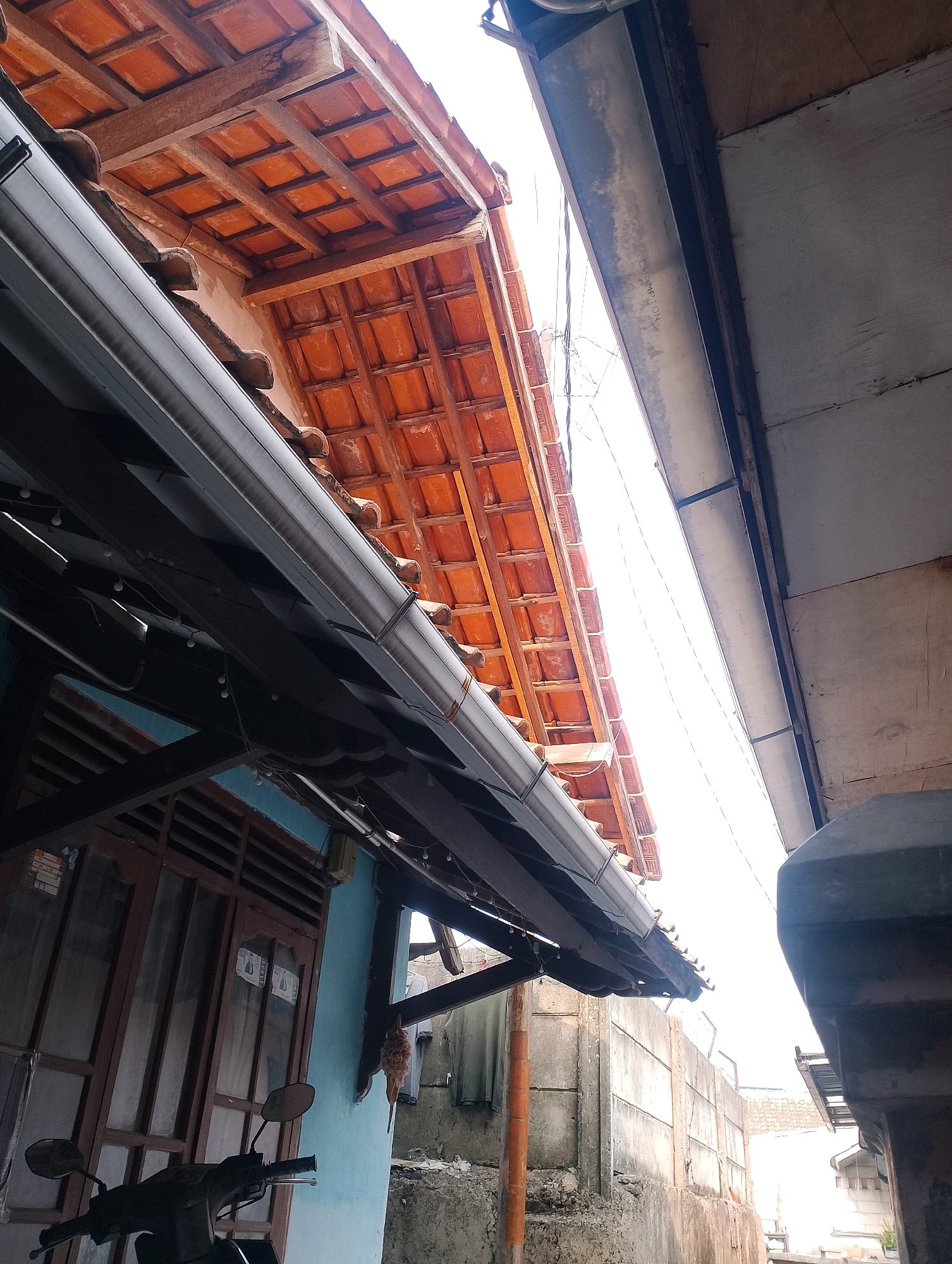 The image shows an outdoor scene in a residential area of Ciomas, Indonesia. The perspective is looking upwards, primarily focusing on the roof structures of adjacent buildings.
In the foreground and mid-ground, a dark grey gutter system runs horizontally across the frame, attached to the underside of a sloped roof. This roof is covered with terracotta tiles and supported by visible wooden beams. To the left of the gutter, a portion of a light blue wall is visible, featuring a wooden-framed window with horizontal slats. A black motorcycle is partially visible in the lower left corner, with its handlebars and mirrors in focus.
To the right of the gutter system, the roofline of another building is seen. This structure appears to have a lighter colored soffit or ceiling paneling, with some text visible, possibly a brand name. Further to the right and in the background, a concrete wall or partition separates areas, with some grey cinder blocks and a section of a building with white patterned siding visible. The overall impression is that of a modest urban or suburban residential environment, possibly an alleyway or narrow street between houses. The sky is bright and washed out, suggesting daylight, but the specific time of day is not determinable from the lighting. There are no people visible in the frame.