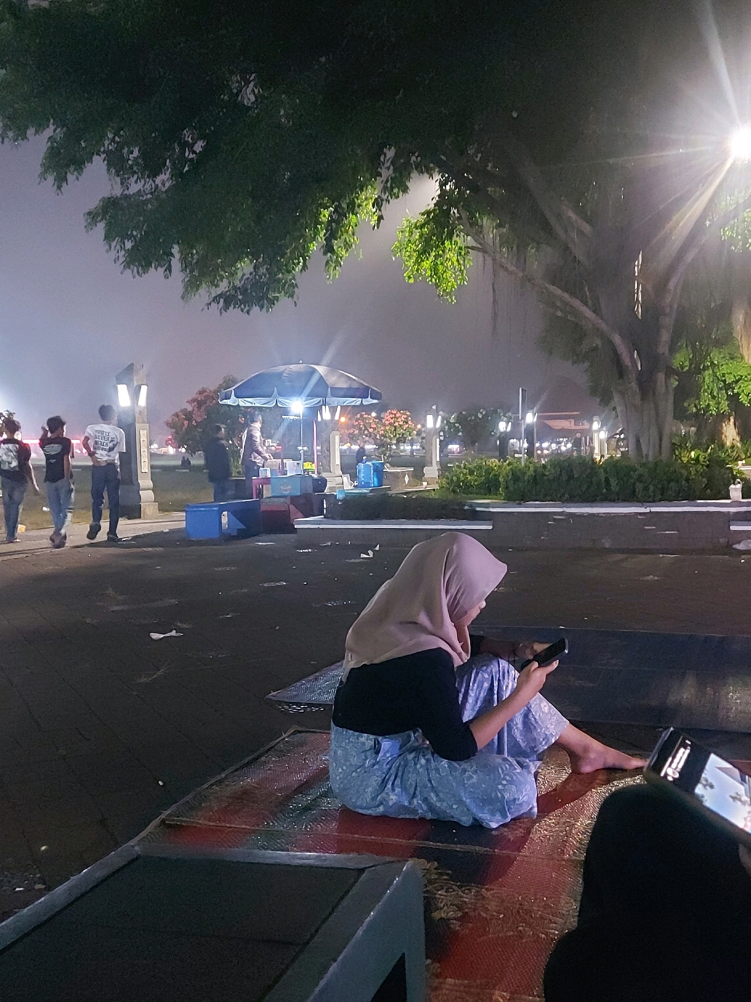 In the city of Wonosobo, Indonesia, at night, a woman wearing a light pink hijab and a black top is seated on a patterned mat, looking at her smartphone. She is wearing light blue patterned pants. Her bare feet are visible. Another person, partially visible and wearing dark clothing, is also seated and looking at a smartphone. The background shows a large tree with hanging roots, illuminated by a bright streetlight. In the middle ground, there are several people, some standing and walking near a food stall under a blue umbrella. Blue and red plastic containers are visible around the stall. The ground is dark and appears wet in places.