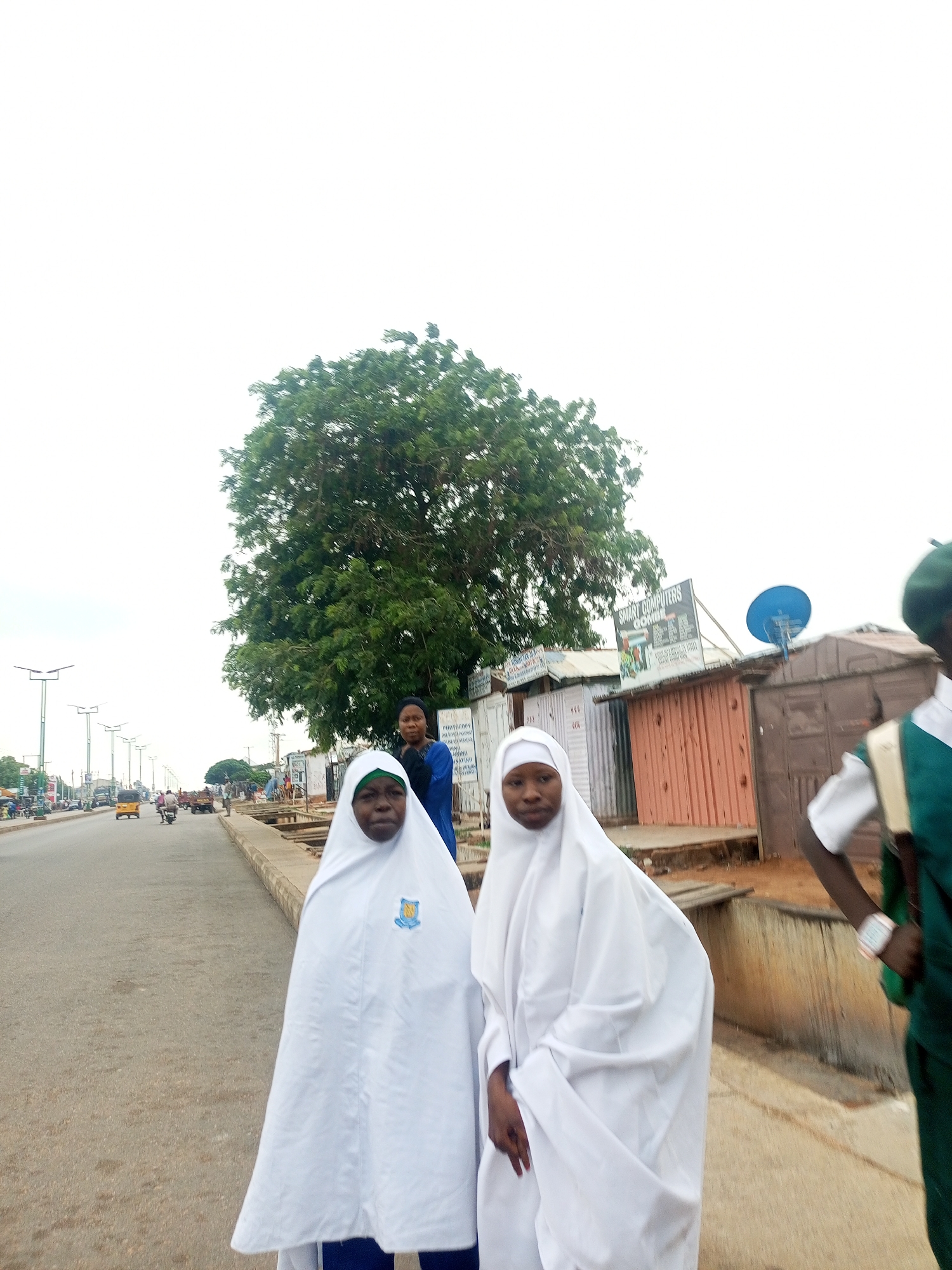 In an outdoor, overcast urban scene, two young females wearing white Islamic hijabs stand on a paved road. The female on the left has a green headscarf peeking out from her white hijab and a school crest visible on her garment. The female on the right is also wearing a white hijab. Behind them, a large tree with green foliage dominates the background, with a street with traffic, buildings, and a satellite dish visible to the right. A person in a green uniform is partially visible on the far right of the frame.