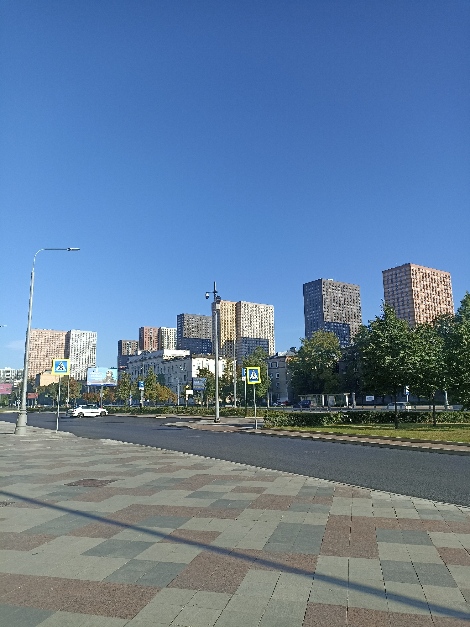 The image depicts an urban scene in Moscow, Russia, under a clear blue sky. In the foreground, a patterned sidewalk made of large square tiles in shades of grey, pinkish-brown, and beige stretches diagonally across the lower portion of the frame. Adjacent to the sidewalk is a newly paved asphalt road.
Further back, a street separates the sidewalk from a green median strip. On the median, there are pedestrian crossing signs with blue backgrounds and white pedestrian symbols. A white car is visible on the road, traveling away from the viewer. Beyond the road, a line of trees with green foliage lines the street.
The background is dominated by a row of tall, multi-story apartment buildings. These buildings vary in color and architectural style, with some exhibiting a cream or light orange facade, others a dark grey, and one a muted greenish-grey. Some of these buildings have visible balconies and windows. On the right side of the frame, a lamppost with a security camera attached is visible, and further to the right, more trees can be seen. Several other lampposts are positioned along the street, including one on the left side of the image with a prominent street light. A large billboard with text and an image of a person is visible behind some of the trees and buildings. The overall impression is a modern urban environment, likely a residential area in Moscow, captured on a bright, sunny day.