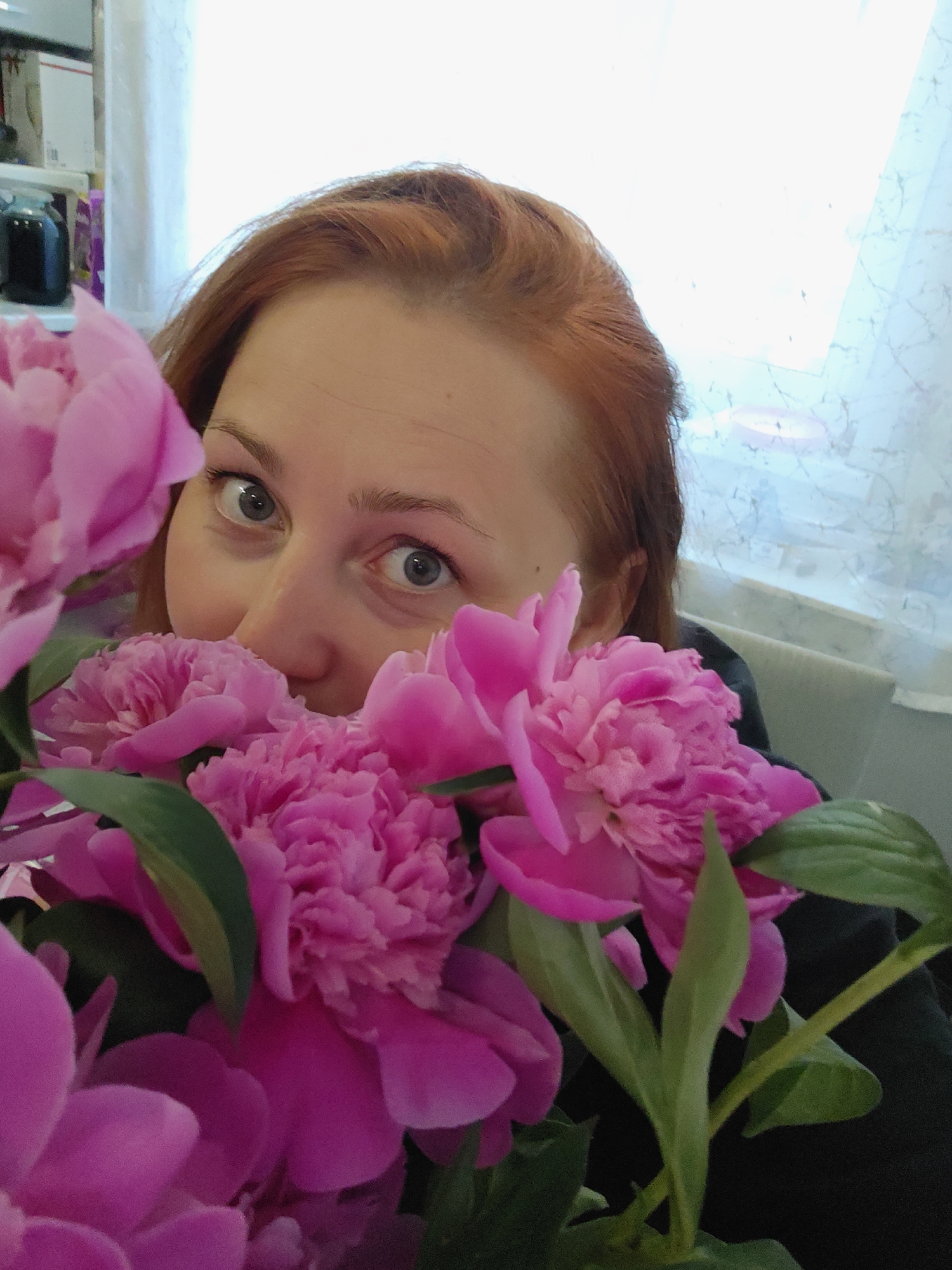 The image is an indoor portrait featuring a woman partially obscured by a large, vibrant bouquet of pink peonies. The woman, with reddish-brown hair, looks directly at the viewer with wide, light-colored (blue/grey) eyes, conveying a playful or curious expression. Only her forehead, eyes, and the upper part of her nose and cheeks are visible above the dense floral arrangement.
The bouquet of peonies, showcasing numerous fully bloomed, rich pink flowers with dark green leaves, dominates the foreground and extends across much of the lower frame, enveloping the woman.
The background is softly lit by natural daylight, indicating daytime. On the right, a sheer white curtain covers a window, allowing bright light into the room. A light-colored chair or piece of furniture is faintly visible behind the woman on the right. On the blurred left side of the background, a shelf holds a dark blue jar or bottle and some white boxes. The scene suggests a cheerful and intimate domestic setting in Markovskii, Russia. No visible text is present in the image.