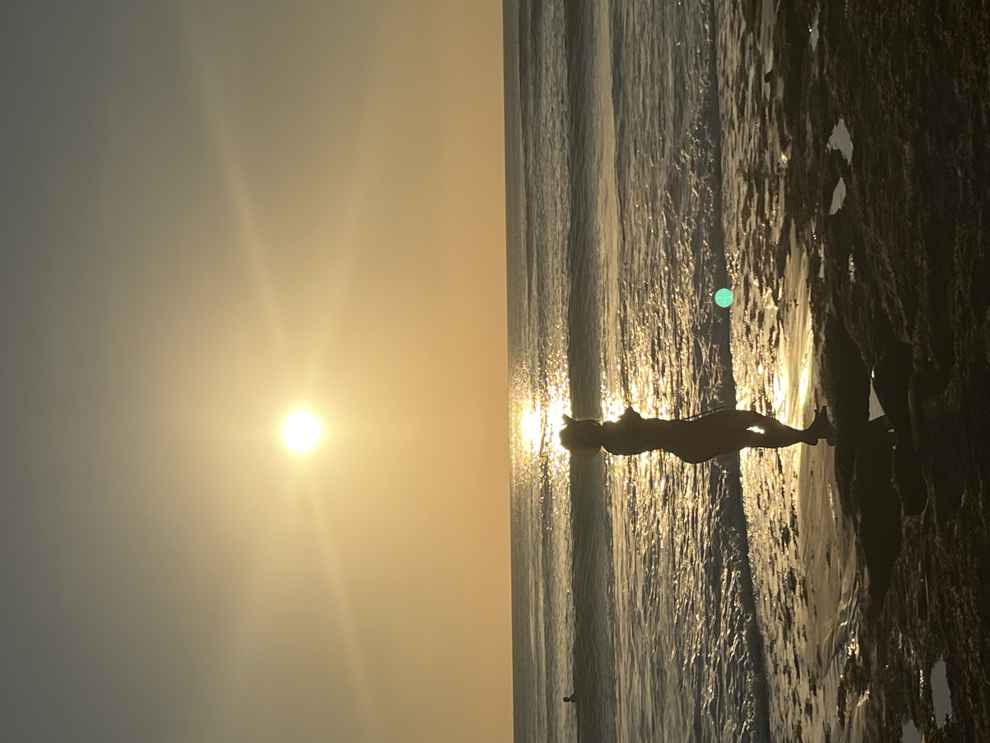 A person wearing a hat and swimwear stands silhouetted on the shore of a beach in Kuta Selatan, Indonesia, facing the ocean during sunset. The sun is low in the sky, casting a warm, golden glow that reflects intensely on the water's surface. Gentle waves lap at the shore, and the sand in the foreground is dark and appears wet in places, with small pools of water reflecting the sky. In the distance, a faint silhouette of another person can be seen in the water. The overall atmosphere is serene and peaceful, characteristic of a sunset on a tropical beach.