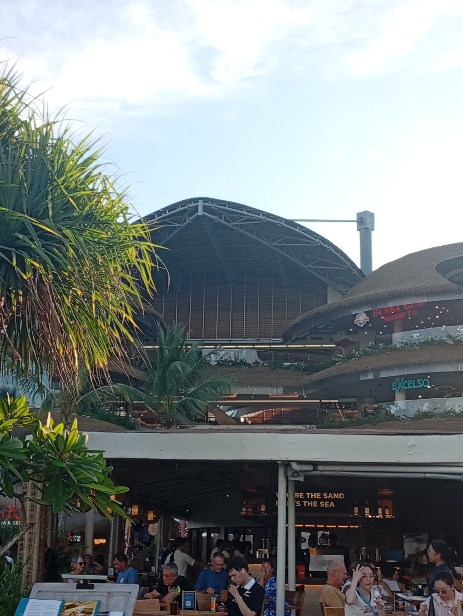 This media file captures a multi-level commercial complex in Denpasar, Indonesia, bustling with activity, likely during the daytime or late afternoon. The scene is characterized by lush tropical foliage, including prominent palm trees and pandan-like leaves, integrated into the architecture.
In the foreground, an outdoor or semi-outdoor restaurant area is filled with people seated at tables, engaged in dining and conversation. They are casually dressed, contributing to a relaxed atmosphere. The building features various levels with open-air designs and thatched roofs.
Visible signage includes "BUBBA GU SHIRMP CO.," which is likely Bubba Gump Shrimp Co., and "EXCELSO," indicating a coffee shop. Another partial sign reads "RE THE SAND 'S THE SEA," suggesting a slogan or theme for one of the establishments. "JA & GRI" is also partially visible, possibly indicating part of a restaurant's name.
In the background, a large, modern building stands with a distinctive arched, industrial-style roof structure, contrasting with the more traditional thatched roofs of the lower levels. The sky above is bright with some scattered clouds. The overall impression is of a lively, recreational area combining modern design with tropical elements.