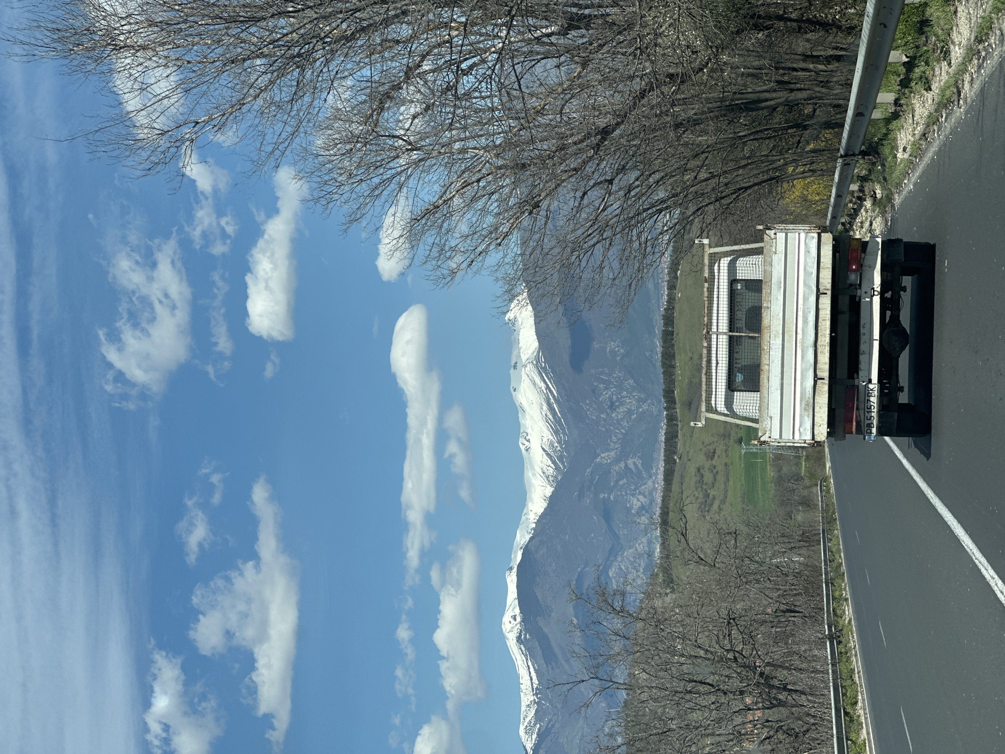 The image depicts a road in Mihiltsi, Bulgaria, with a white truck in the foreground, partially obscuring the view. The truck's license plate number is PB5157BK. In the background, a mountain range with snow-capped peaks rises. The sky is blue with scattered white clouds. Bare trees line the side of the road. The road has a white line marking the lanes. A metal guardrail is visible along the roadside. A green field is present in the mid-ground between the road and the mountains.