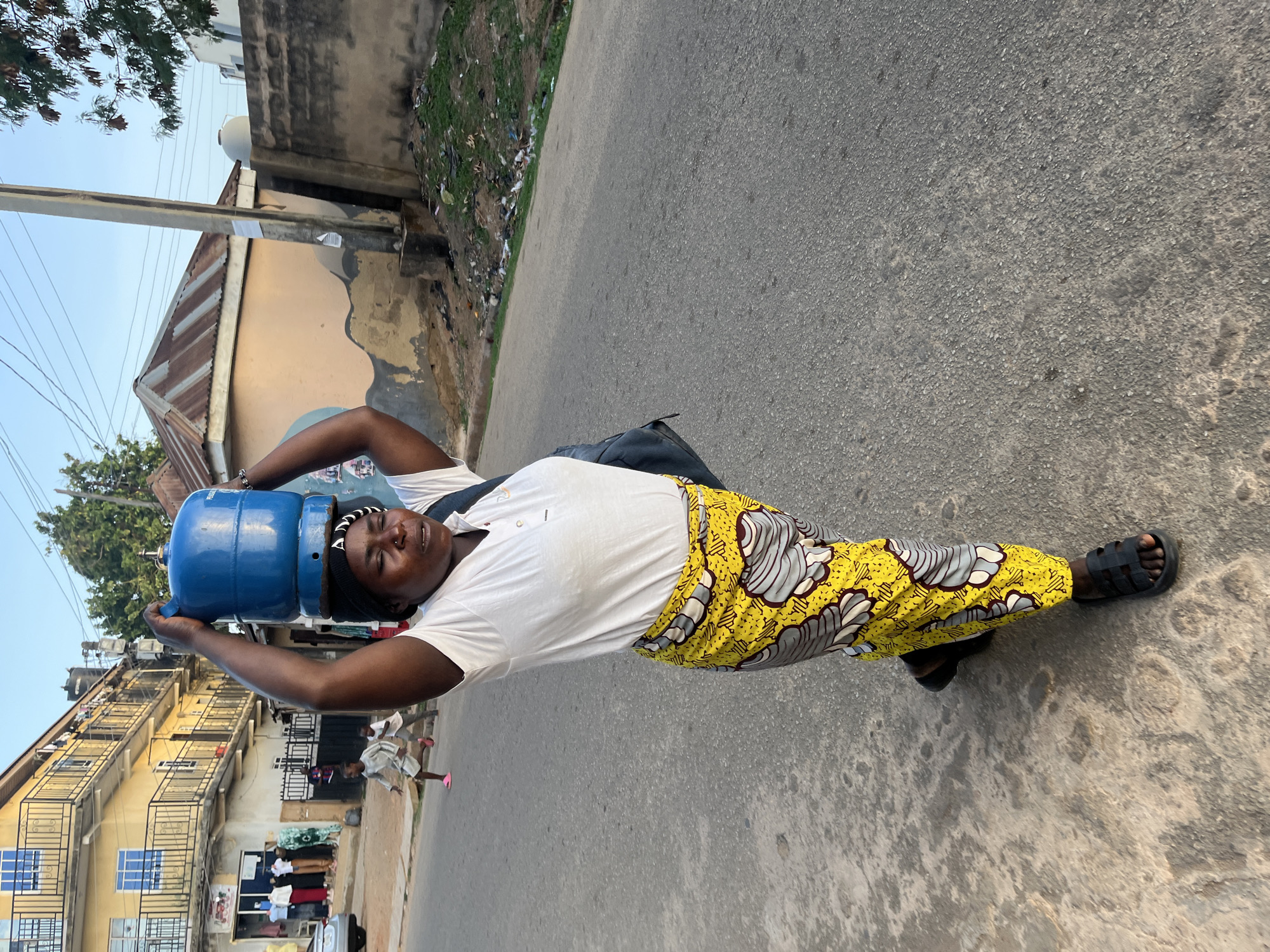 A woman stands on an asphalt street in Jos, Nigeria, balancing two blue gas cylinders on her head. She wears a white t-shirt, yellow patterned pants, and black sandals. A black bag hangs over her shoulder. Buildings line the street on either side, and litter is visible along the curb. The sky is visible in the background.