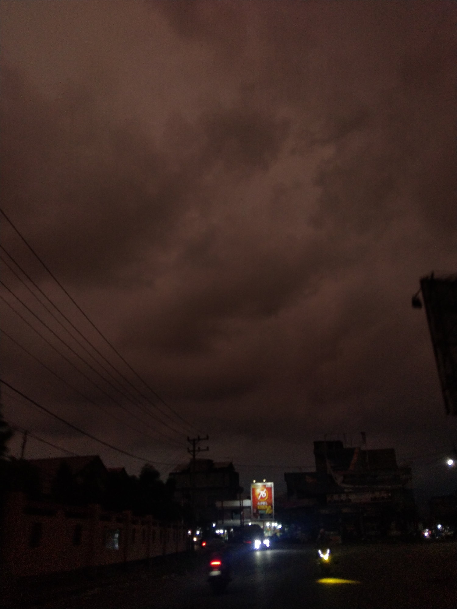 The image depicts a nighttime scene in Meukokuthang, Indonesia. The sky is overcast with dark, heavy clouds. In the foreground, a road is visible with vehicles, including a motorcycle with its headlights on. Along the side of the road are buildings, some of which appear to be residential, and a utility pole with power lines. A billboard with the number "76" is illuminated amidst the buildings. The overall environment suggests a dimly lit urban area during a cloudy night.
