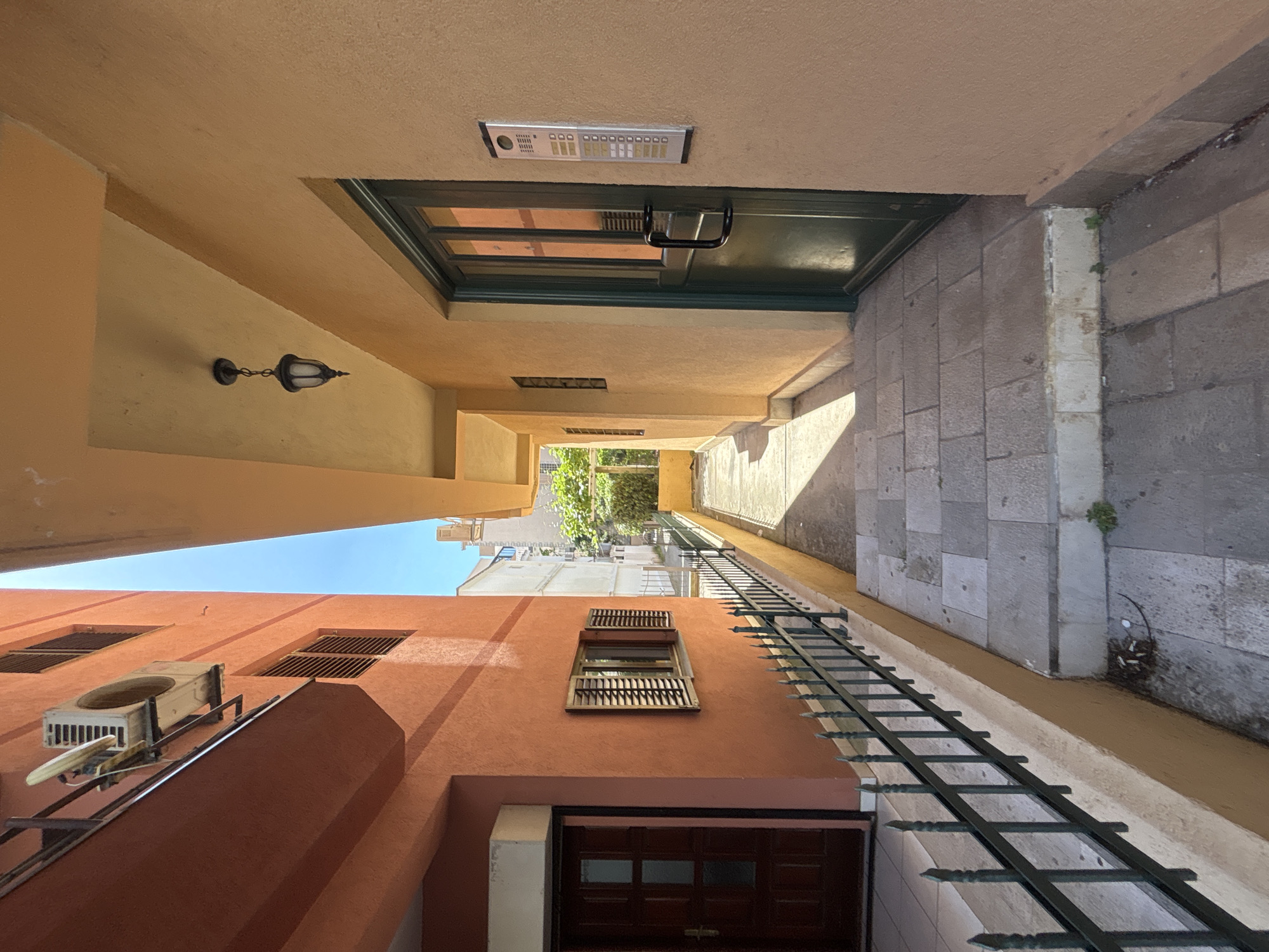 The image depicts an alleyway in the city of Budva, Montenegro. The view is upward, showing the facades of two buildings and the passage between them. One building is painted in shades of orange, with several windows fitted with wooden shutters, an air conditioning unit on an external platform. The other building is painted in yellow. Above the alleyway is a door with a silver intercom system above it. A light fixture is visible on the side of the yellow building. The pavement is tiled. The alleyway leads toward an open space with sunlight and greenery. A dark metal railing runs along the side of the alley.