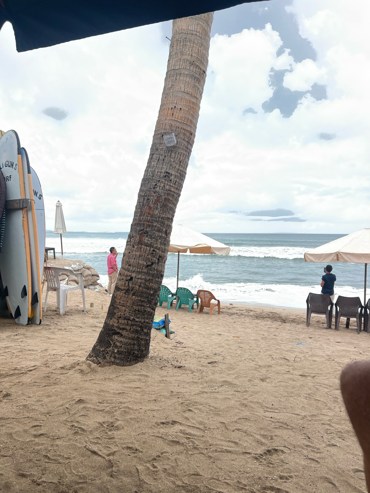 A beach scene in Kuta, Indonesia, shows a sandy shore with the ocean in the background. A large palm tree trunk is prominently featured in the foreground, leaning to the left. Several surfboards are propped up against a wooden structure to the left of the palm tree. In the middle ground, a person in a pink shirt stands near sandbags and a white plastic chair. Further right, two white beach umbrellas are set up, with a person in a dark shirt standing near the right umbrella, looking towards the ocean. Several colorful plastic chairs are scattered on the sand. The ocean has moderate waves and the sky is overcast with clouds.