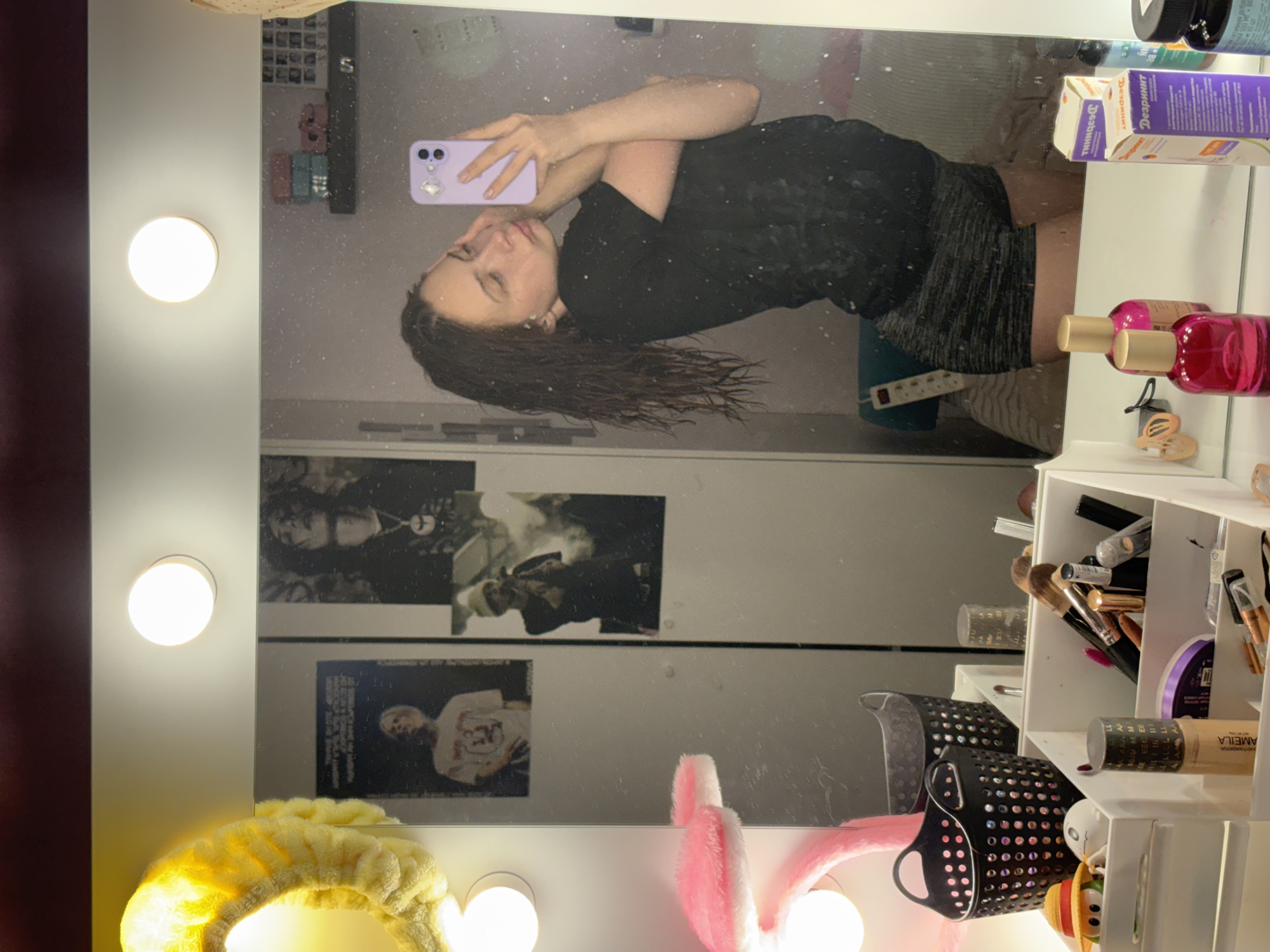 A person with wet, dark hair is taking a selfie in a mirror. The person is wearing a dark, textured top and a dark skirt or shorts. The phone is a light purple color. The mirror is surrounded by lights and has two black and white posters visible. In the foreground, there are beauty products, including brushes, foundation, and spray bottles. The location cannot be determined to be Прикамский, Russia based on the given information.