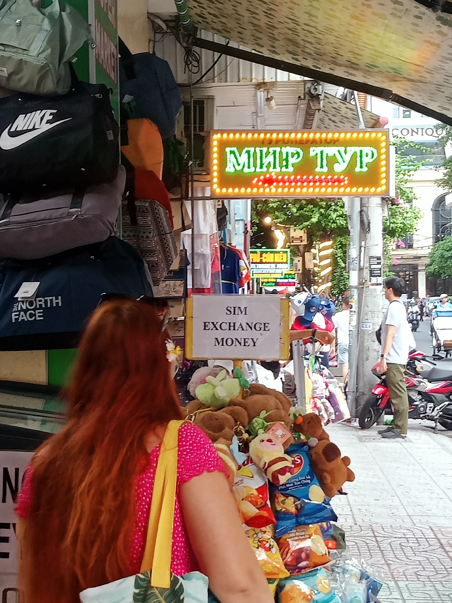 The image shows a street scene in Nha Trang, Vietnam, with a storefront advertising "SIM EXCHANGE MONEY" and a brightly lit sign reading "МИР ТУР" (World Tour). The shop displays items such as bags (Nike, North Face), plush toys, and snacks. A woman with long red hair is standing in front of the store, and a man is visible in the background near red scooters on the sidewalk. There is a business named "CONTOUR" in the background. A variety of people and vehicles are seen in the distant street.