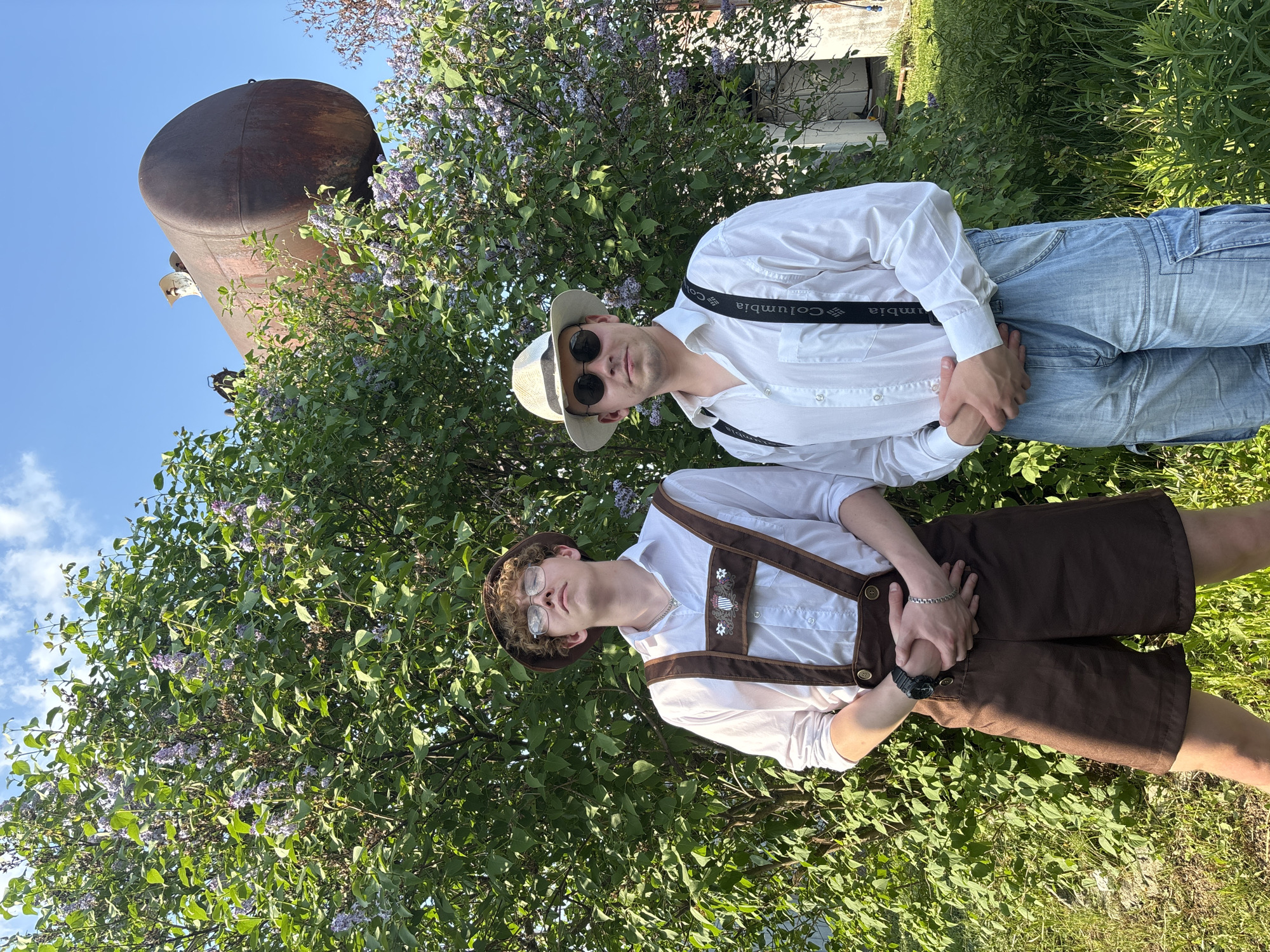 Two young men are standing in an outdoor setting amidst greenery and flowering bushes. The man on the left wears a white button-down shirt, light blue jeans, black suspenders, a light-colored hat, and sunglasses. The suspenders have "Columbia" printed on them. The man on the right wears a white button-down shirt, brown lederhosen, and suspenders with traditional decorative elements. He is wearing glasses and a brown hat. Their hands are clasped together. Behind them is dense foliage with lilac-colored flowers. To the left, a large rusty tank is visible against a blue sky. A small building is partially visible behind the foliage. The location is Пальники, Russia.