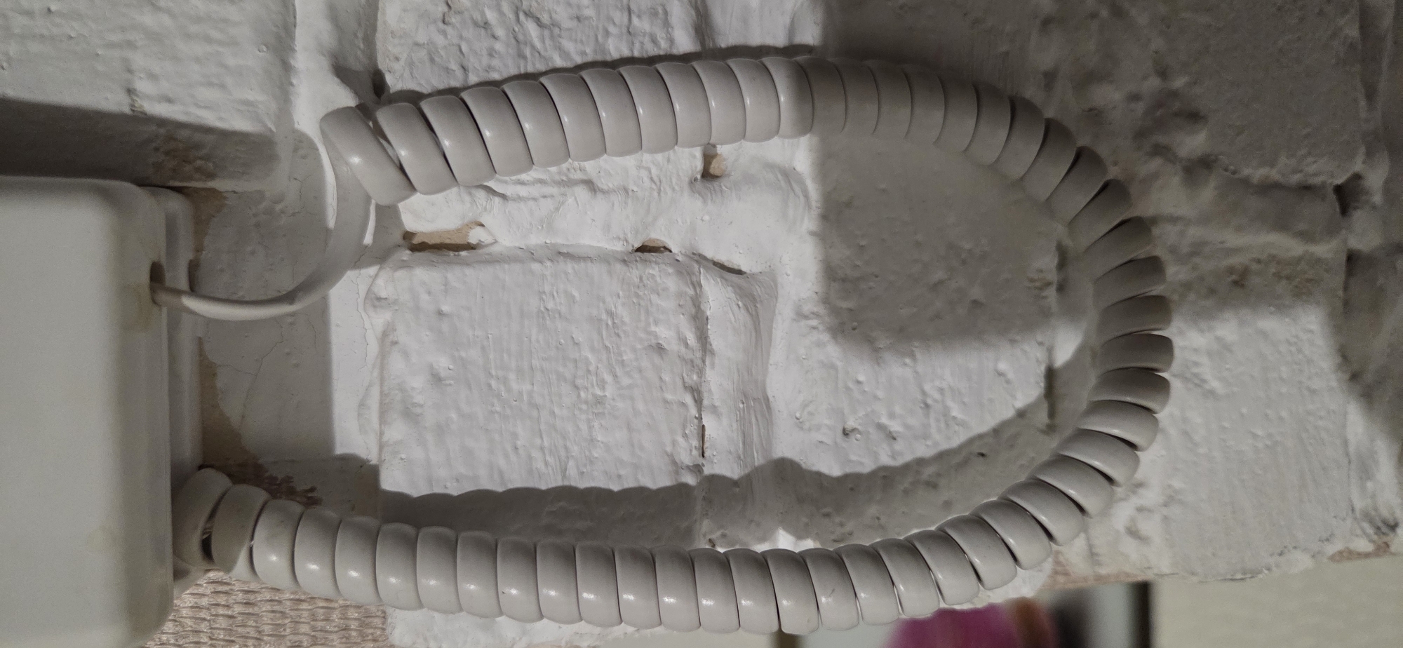 A coiled telephone cord hangs in front of a textured, painted brick wall. The cord is a light gray color and is attached to a cream-colored device mounted on the wall. The wall itself appears to be made of brick and is covered in a thick layer of white paint. The paint is unevenly applied, creating a rough, textured surface with visible brush strokes and some areas where the brick underneath is showing through. The lighting in the image casts a shadow of the coiled cord onto the wall. The overall scene suggests an indoor setting, possibly a home or office. The location is identified as Lipetsk, Russia.