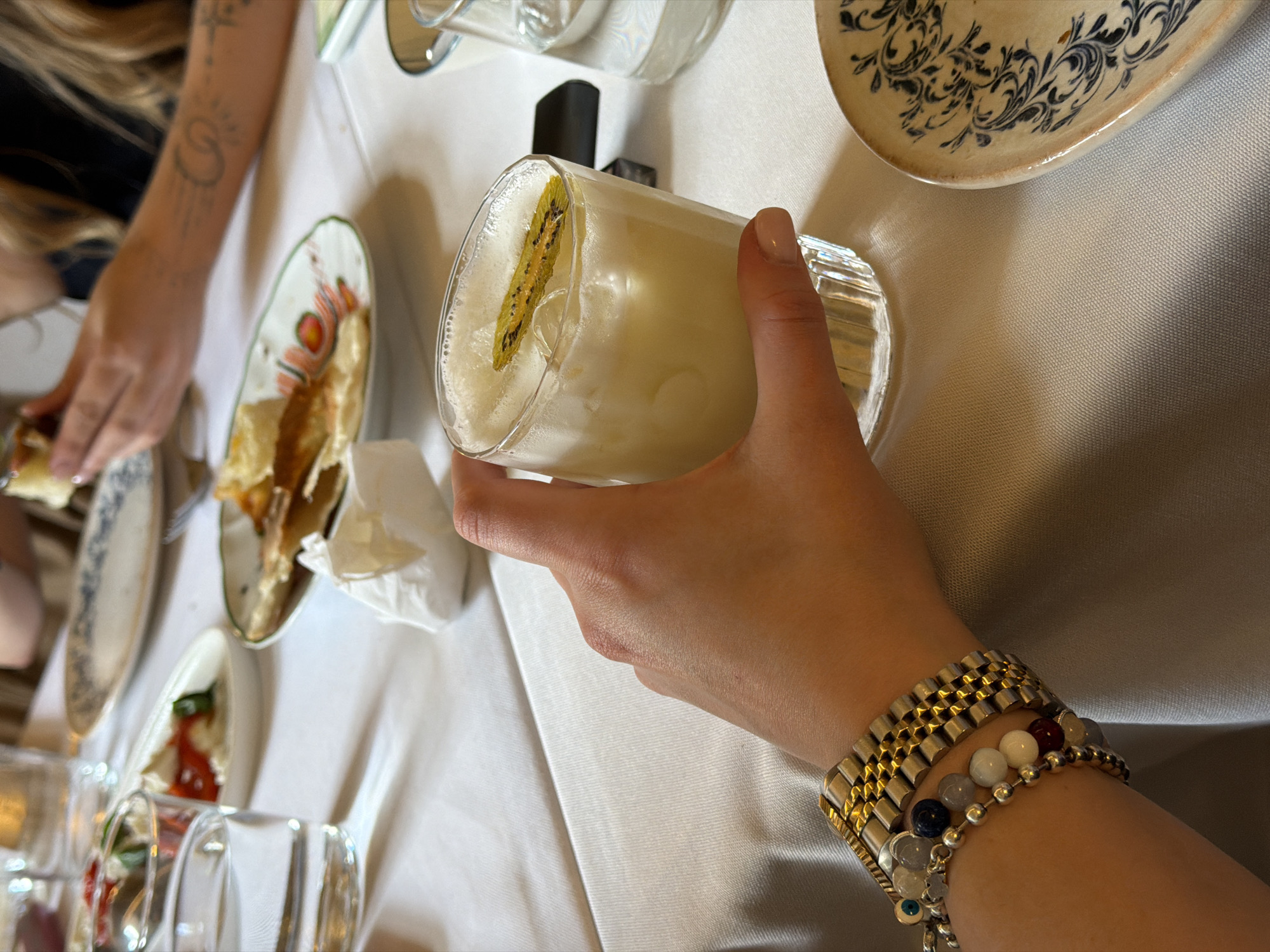 A close-up shot captures a hand holding a cocktail garnished with a slice of kiwi. The drink is in a fluted glass filled with ice and a frothy, pale yellow liquid. Multiple bracelets adorn the wrist of the hand, including a gold watch band, a bracelet with various beads, and another with a blue and white evil eye charm. The scene is set on a white tablecloth, with glimpses of plates and other glasses suggesting a dining or social setting. In the blurred background, a person's arm with tattoos is visible, as is a portion of a decorative plate with a blue and white floral pattern. The overall impression is a candid moment at a restaurant or bar, likely in an urban environment, though the specific city or location in Kyiv, Ukraine, cannot be determined from the visual information alone.