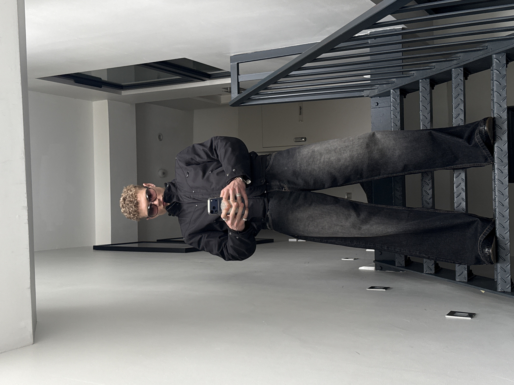 The image shows a person with curly hair, wearing sunglasses, a black jacket, and dark jeans, holding a smartphone and standing in a modern, sparsely decorated interior. The location appears to be a gallery or exhibition space, characterized by white walls and floors. In the background, there's a metal structure with horizontal bars, possibly part of an installation or architectural feature. There are also some small, rectangular objects scattered on the floor. The image shows an indoor scene.
The location in London is not specified.