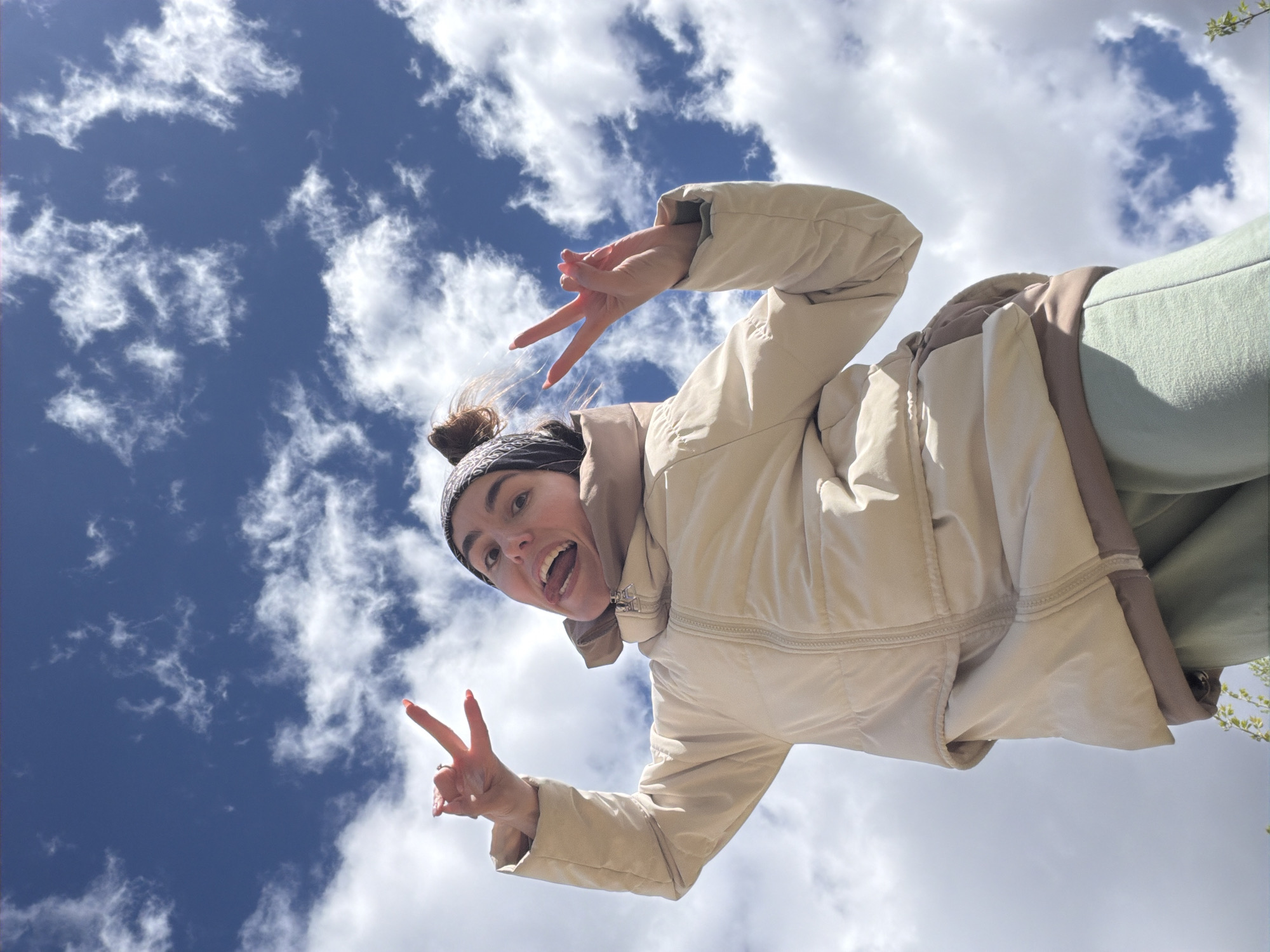 A young woman is captured in a low-angle shot against a backdrop of a blue sky dotted with fluffy white clouds. She is wearing a beige puffer jacket over what appears to be a green tracksuit, and a dark-colored headband holds back her hair, which is styled in a bun. She is making peace signs with both hands and has a wide smile, conveying a sense of joy and excitement. The setting is outdoors, with hints of trees in the lower right corner, but the specific location in Nizhnii Vishur, Russia, is not discernible from the image itself.