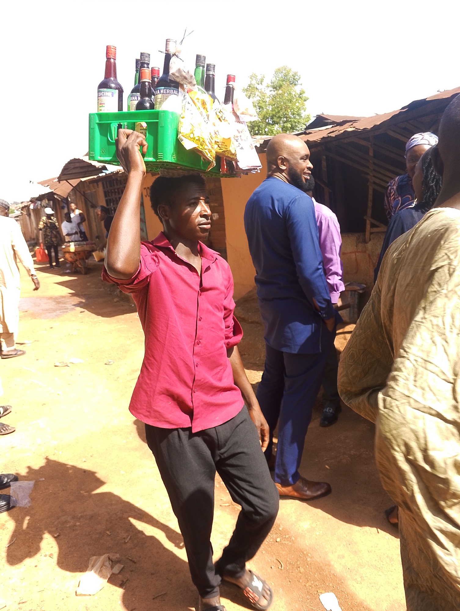 A man in a red shirt and black pants carries a green plastic crate filled with bottles on his head. The bottles appear to be various herbal medicines and beverages. The man is walking on a dirt road, seemingly selling the items. In the background, there are several other people, some of whom appear to be engaged in conversation. Buildings with corrugated metal roofs are visible in the background, suggesting a market or commercial area in Jos, Nigeria. The lighting indicates it is daytime.