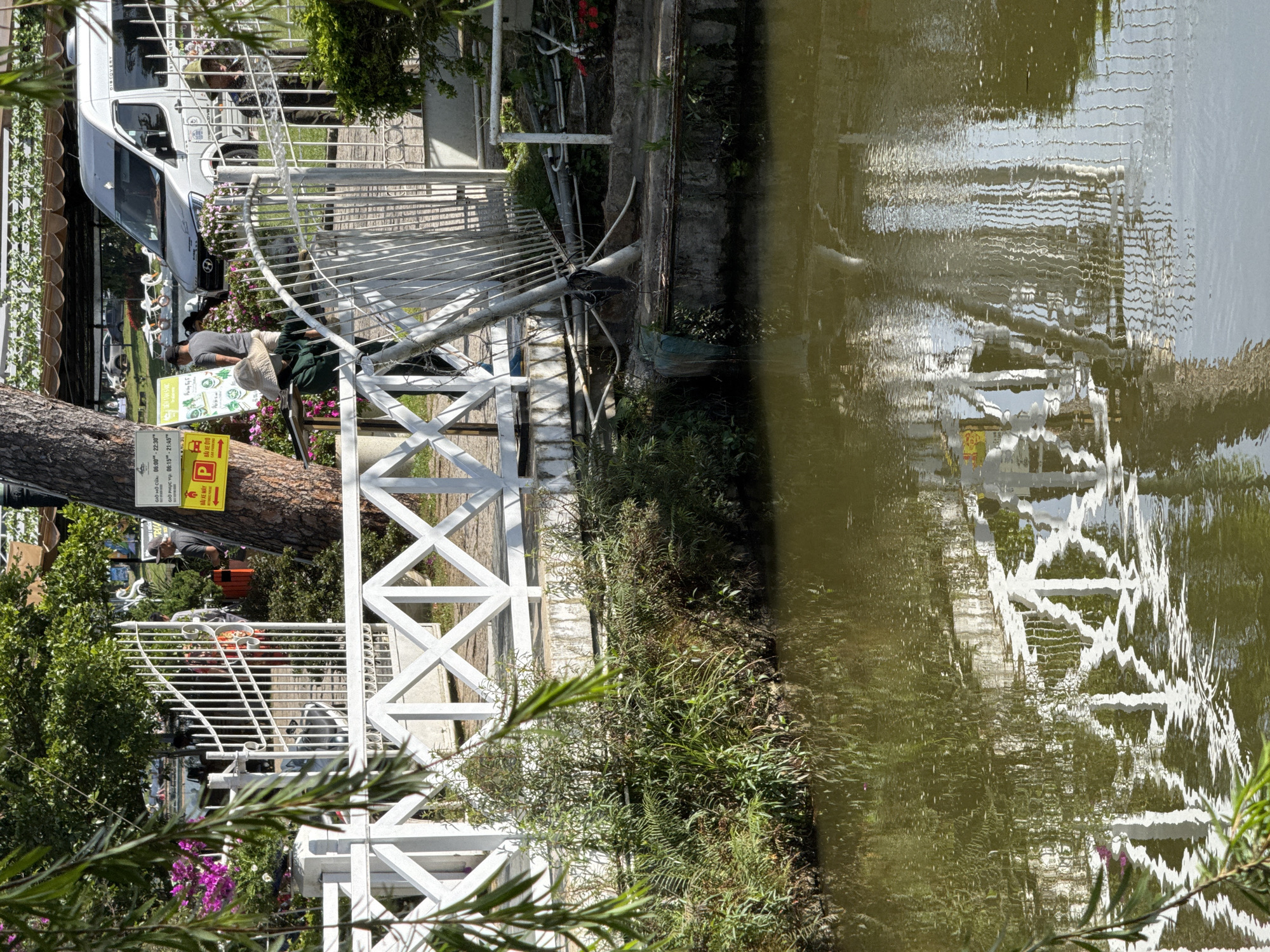 The image depicts a canal in Da Lat, Vietnam, with a white wooden bridge spanning across it. The bridge is adorned with ornate railings and posts. On the left side of the canal, lush green foliage grows along the bank, with a few pink flowers visible. Beyond the bridge, a white van is parked, and several people are present, some tending to plants and others appearing to be customers. A signpost with yellow and red lettering is attached to a tree near the bridge. The water in the canal reflects the bridge, trees, and surrounding structures, creating a distorted yet recognizable image. The scene appears to be a public space, possibly a garden or a market, given the presence of plants, a van, and multiple individuals.