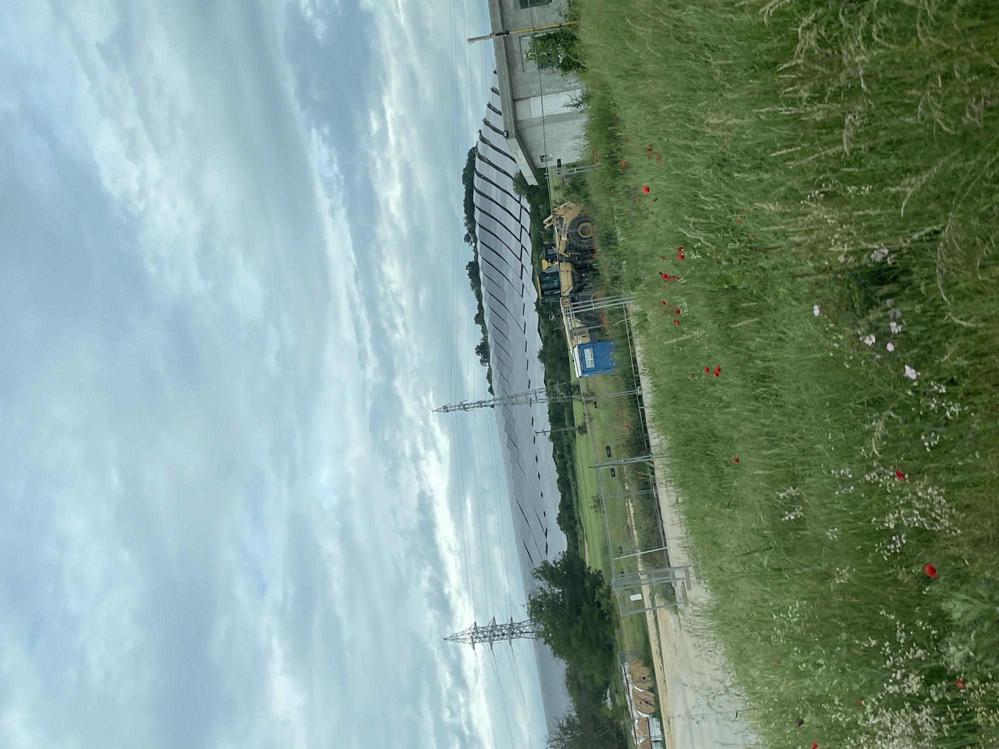The scene depicts a large solar panel array located near Kamenar, Bulgaria. The panels are arranged in rows and stretch across a grassy field. Adjacent to the panels, there is heavy machinery, likely used for maintenance or construction. A tall, metal transmission tower is also visible, suggesting the connection to the electrical grid. The foreground features overgrown vegetation with numerous wildflowers. The sky is overcast, indicating cloudy weather conditions.
