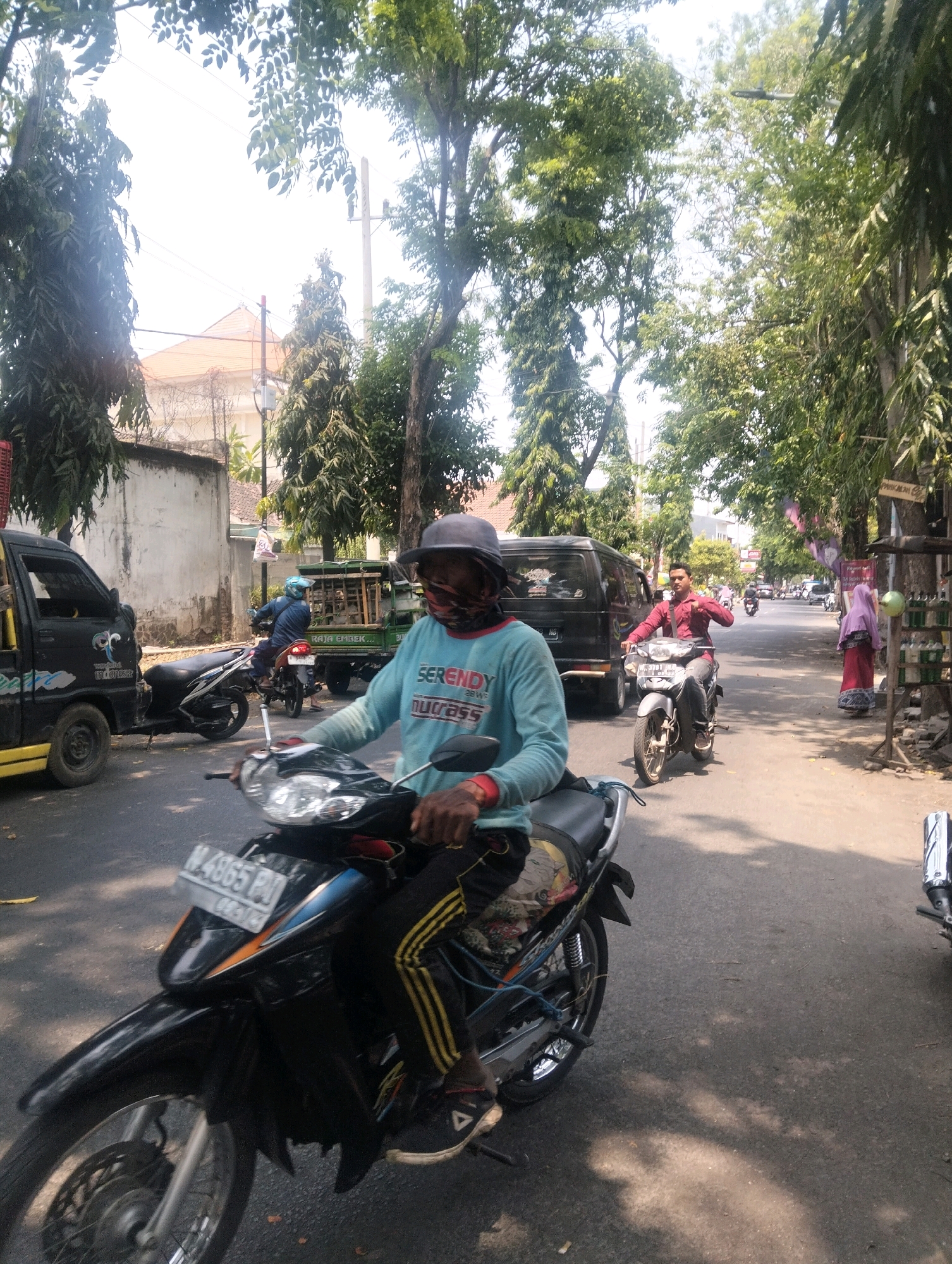 The image depicts a street scene in Semampir, Indonesia. In the foreground, a person wearing a blue long-sleeved shirt, a face mask, and a grey hat is riding a black motorcycle. They are wearing black pants with yellow stripes and dark athletic shoes. The motorcycle has a license plate visible with the characters "N 4865 PN". To the left, a black truck is parked. In the middle ground, another person is riding a silver motorcycle, wearing a red long-sleeved shirt and dark pants. Behind them, a green truck is also present. Further down the street, a woman in a pink hijab and red dress is standing next to a stall displaying various bottles. The street is lined with trees, and buildings are visible in the background under a bright sky. The overall atmosphere suggests a typical day in an Indonesian urban environment.