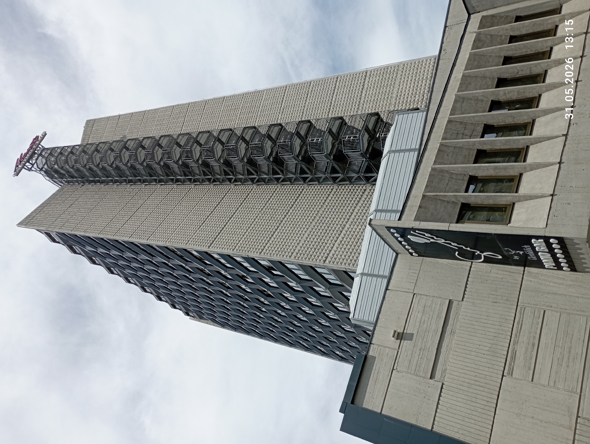 An upward angle shot shows a high-rise building in Burgas, Bulgaria. The building is constructed from concrete slabs with textured triangle patterned walls. A fire escape extends upwards from the building. At the top of the fire escape are the words "Hotel Burgas." The lower half of the building has darker, patterned panels. A concrete structure is in front of the high-rise, partially obscuring it. On the right side of this concrete structure is a sign for a piano bar by Undefeated. The sky is visible behind the buildings. The date "31.05.2026 13:15" is stamped in the upper right corner of the image.