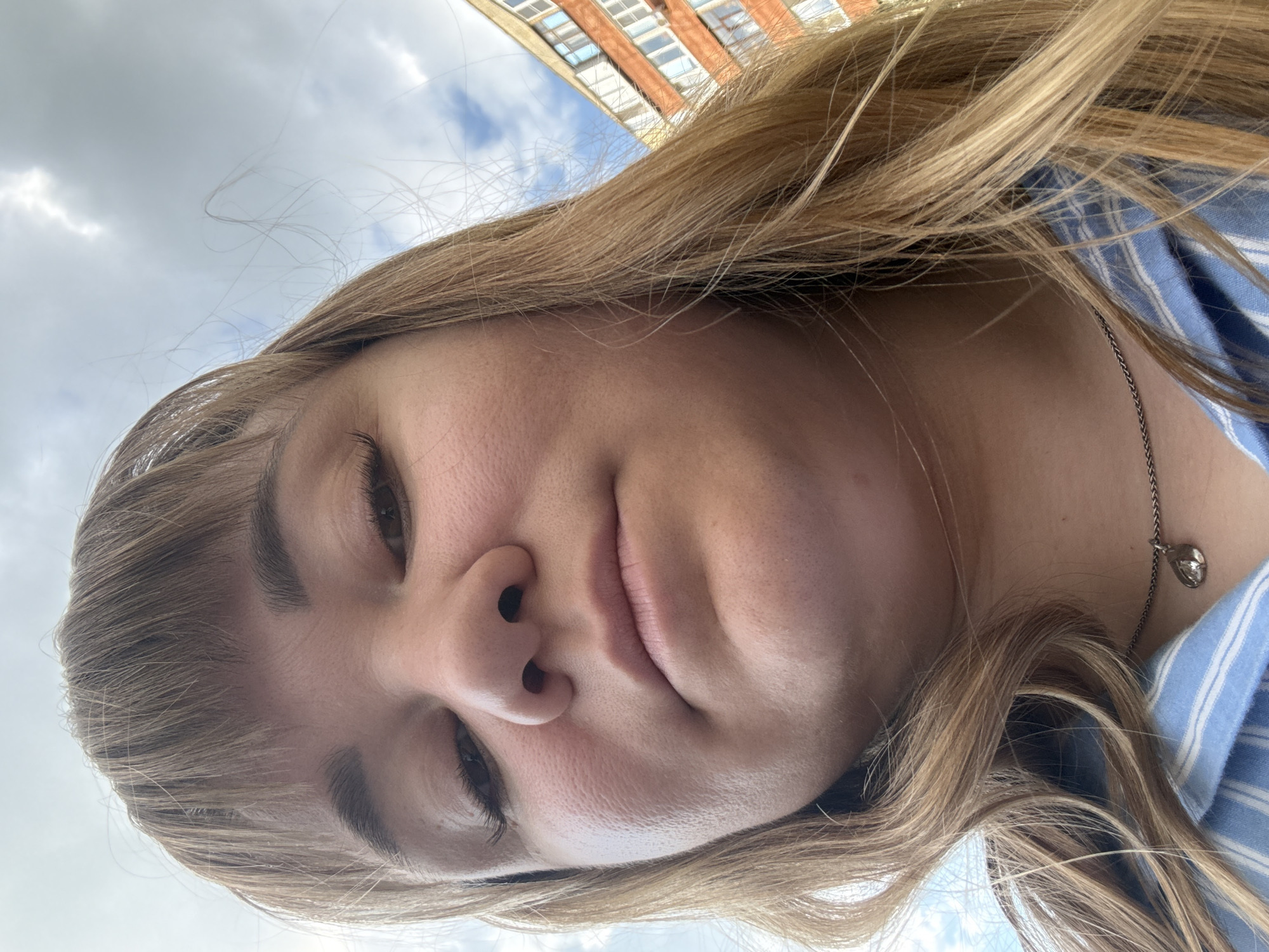 This media file is a close-up, tilted selfie of a young person outdoors. The subject has blonde hair and is wearing a blue and white striped shirt, along with a silver necklace with a small pendant. The focus is on the subject's face, with their eyes looking forward. The background includes a cloudy sky and the upper floors of a brick building, suggesting an urban environment. The lighting and cloudy sky suggest it might be daytime, possibly overcast. The image is oriented sideways. There is no visible text.