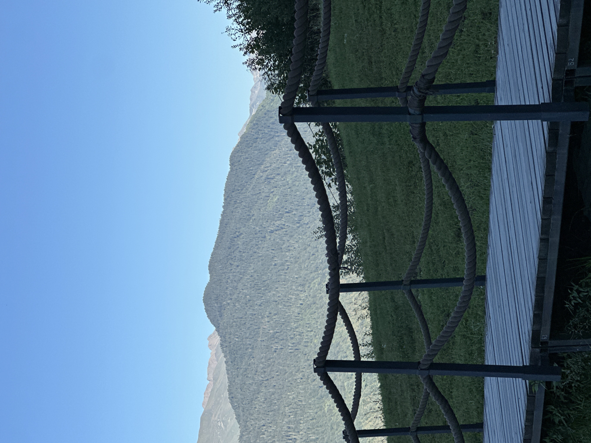 A wooden walkway with a rope railing is in the foreground, leading towards a grassy slope. In the mid-ground, a dense forest covers a mountain. The background features a clear blue sky and distant mountain peaks. The image appears to be taken in Estosadok, Russia, given the presence of the wooden walkway with numbered markers (52, 63) which are common in tourist areas. There are no people visible in the image. The scene is serene and showcases a natural mountain landscape.