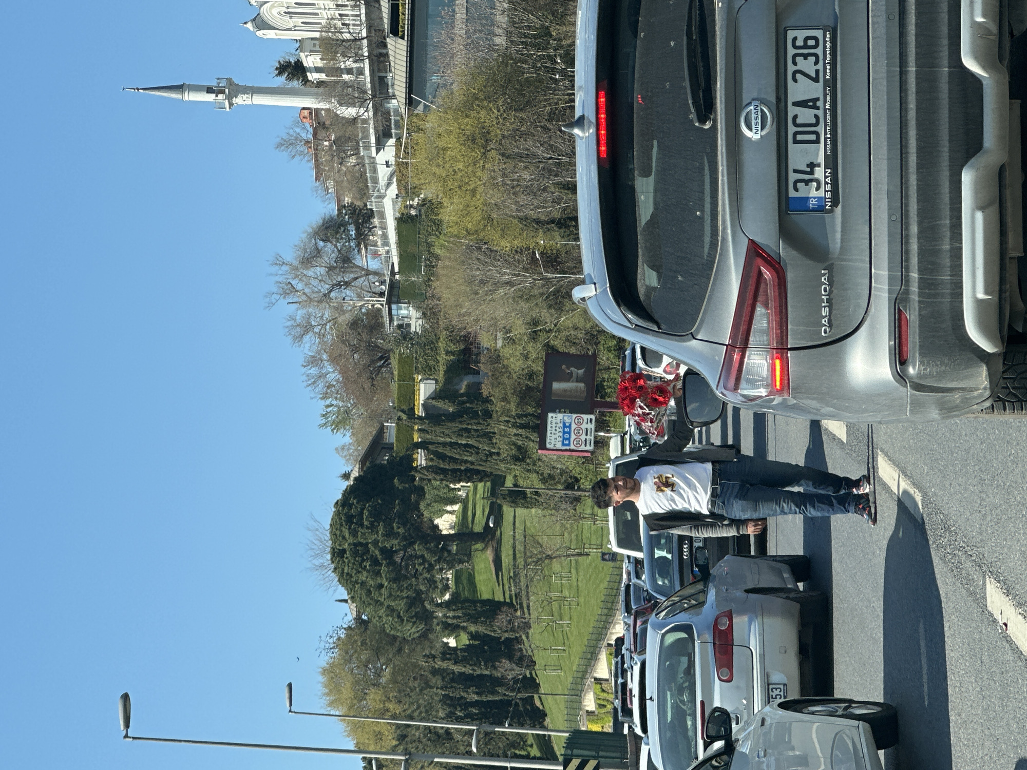 The image depicts a traffic scene in Üsküdar, Turkey. A man is standing between stopped cars, holding a bundle of red flowers, seemingly offering them for sale. He is wearing jeans, a white t-shirt, and a black jacket. Behind him is a gas station. A gray Nissan Qashqai with the license plate "34 DCA 236" is visible in the foreground. Across the street, a park with trees and manicured lawns can be seen. In the background, a white building with a minaret, typical of Turkish mosques, is visible. The sky is clear and blue. The cars are tightly packed, suggesting heavy traffic.