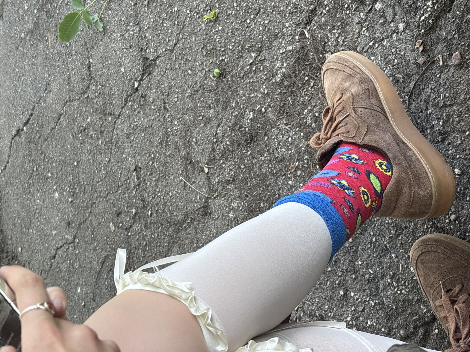 A photograph shows a person sitting on asphalt, presumably in the city of Sofia, Bulgaria, as this information was given, with their legs extended. The visible leg is adorned with a white stocking that has a ruffled edge. Above the stocking, there is a red sock with a pattern of blue, yellow, and green motifs, topped with a blue band. The person is wearing a brown suede sneaker. Another brown sneaker is partially visible. The person's hand is holding a cell phone and wearing a ring on one of their fingers. The asphalt surface is visible in the background, showing signs of wear and containing small stones or gravel. A small plant is also present.