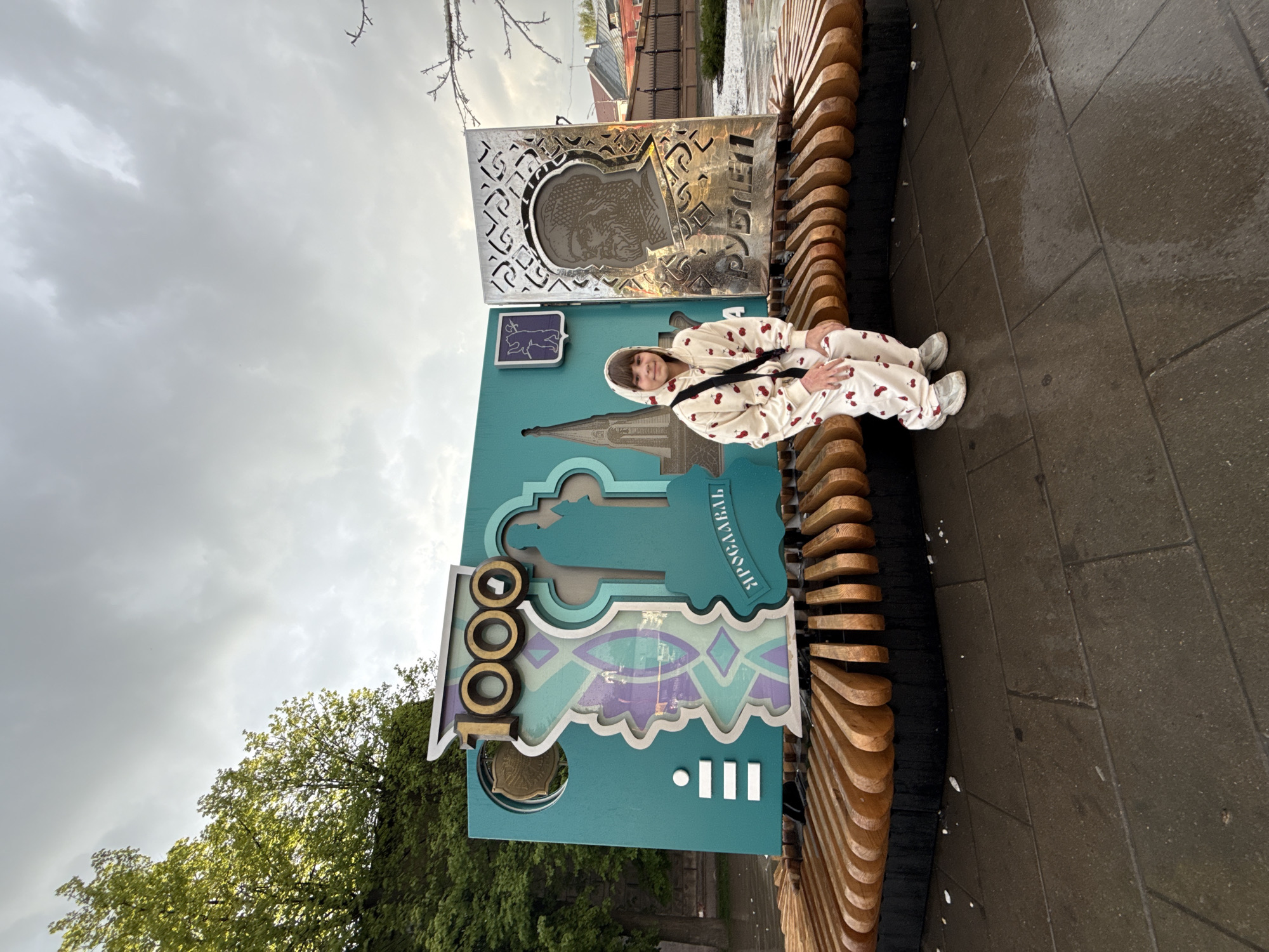A full-body shot shows a person in light-colored pajamas sitting on a bench next to an ornate, teal-colored sign. The bench is made of curved wooden slats connected to a black structure. The sign is decorated with architectural motifs and Cyrillic script. Above the teal sign is a silver-colored plaque with a cut-out design. The ground is made of dark, wet pavement. In the background, trees are visible on one side and buildings on the other. The sky is overcast. The scene is in the city of Заволжье, Russia.