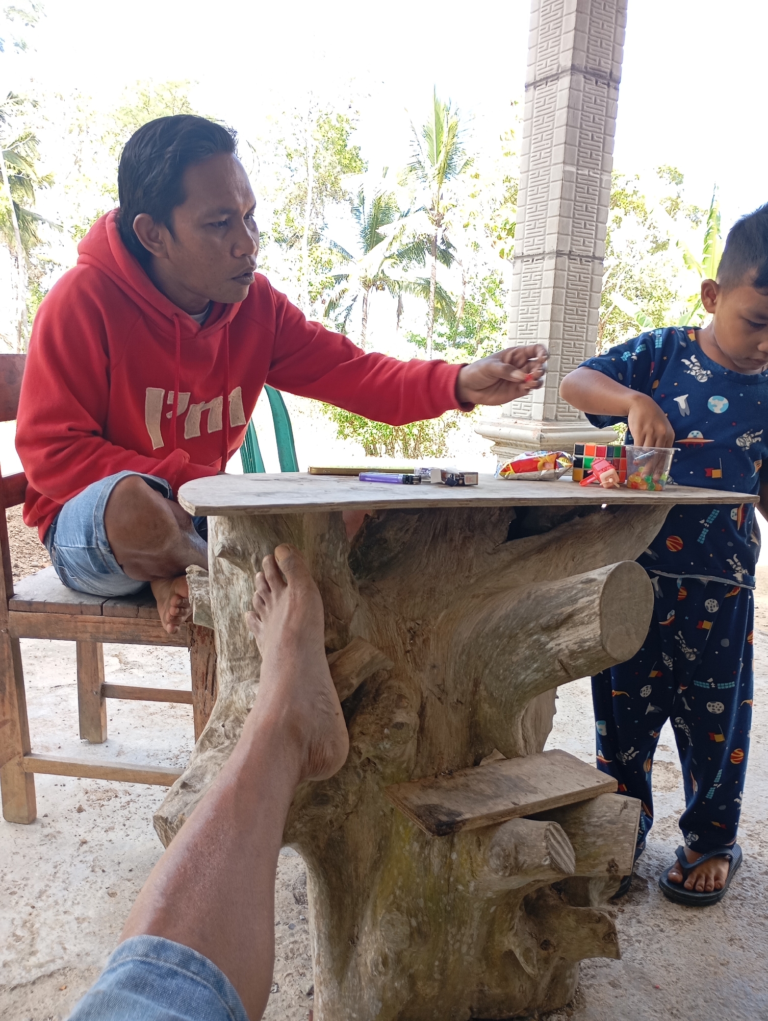 In Ketepung, Indonesia, two individuals are engaged in an activity at a rustic wooden table. A man, wearing a red hooded sweatshirt and denim shorts, is seated on a wooden chair. He is reaching out a hand, holding a small red object. Beside him, a young boy in a blue pajama set with a space-themed pattern stands near the table, his attention focused on items on the table. The table itself is uniquely crafted from a large piece of wood, its surface uneven and textured. Scattered on the table are various objects including a bag of snacks, a Rubik's cube, a small toy figurine, and a clear plastic cup filled with colorful small balls. In the foreground, a bare leg, presumably belonging to the photographer, is visible, resting near the table. The setting appears to be outdoors, with lush green foliage and palm trees visible in the background, suggesting a tropical environment. The ground is a concrete surface.